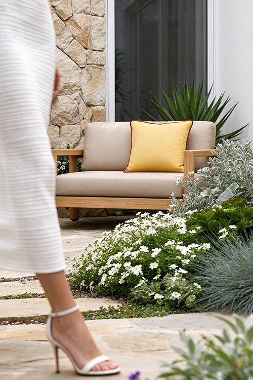 A patio scene with a beige cushioned outdoor sofa, a yellow pillow, and surrounding greenery including white flowers and large plants. A woman wearing a white dress and high heels is walking past.