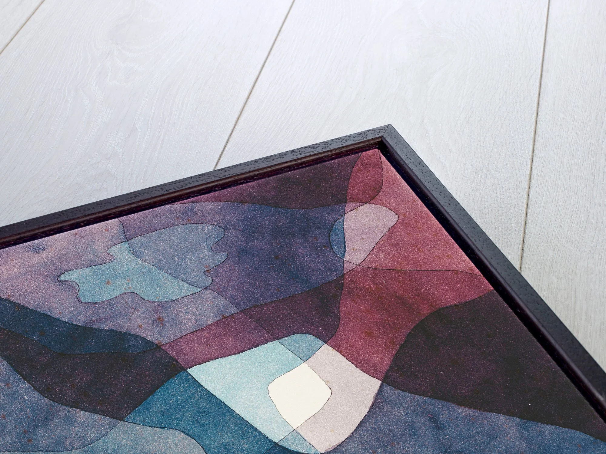 Close-up of a colorful, abstract artwork with various shades of blue, pink, and purple in a black frame, resting on a white wooden floor.