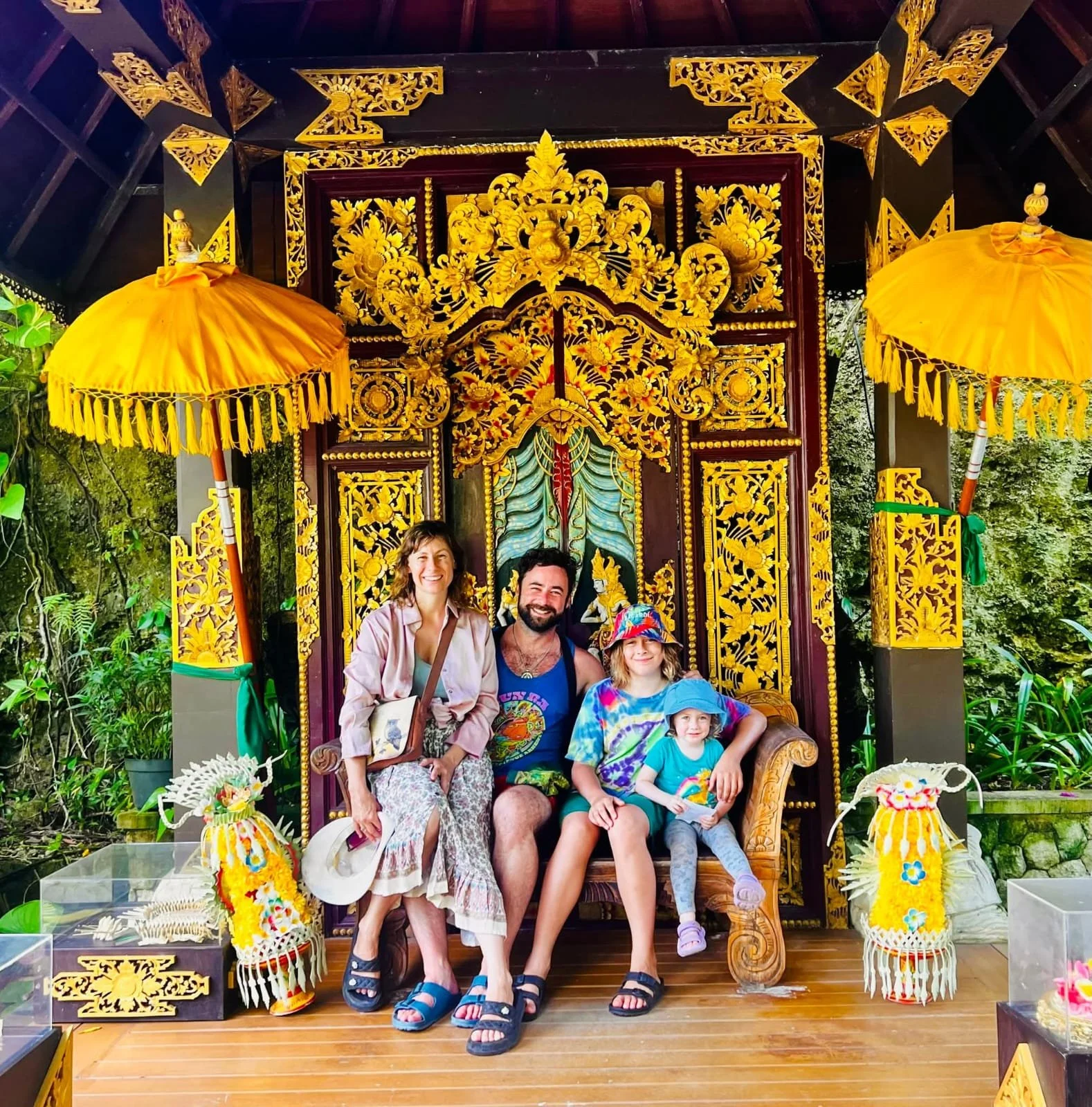 A family of five sitting on an ornate wooden throne with intricate gold carvings, flanked by yellow umbrellas and decorative flowers, outdoors surrounded by greenery.