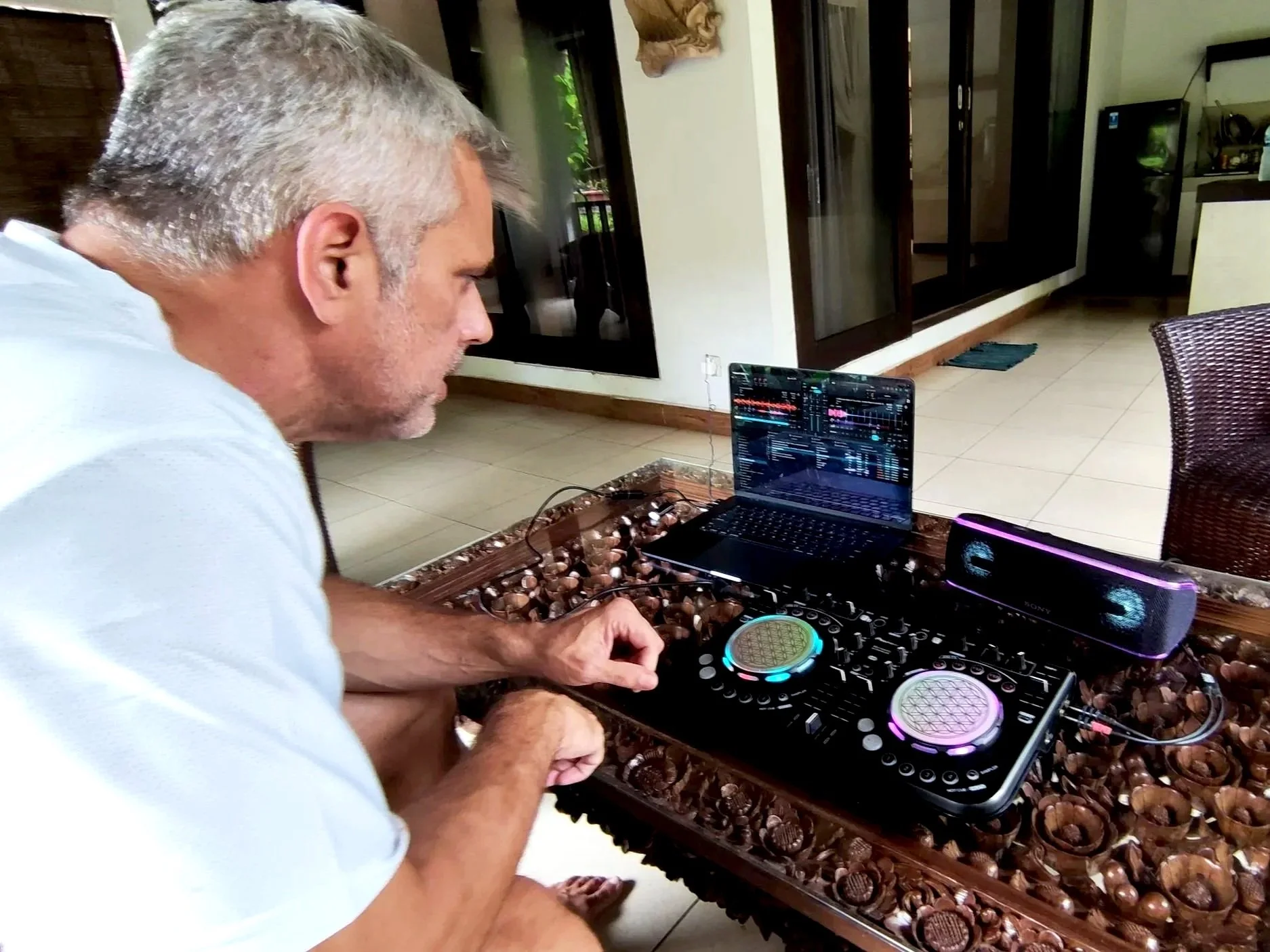 A man with gray hair is DJing at a table filled with chocolates, using a DJ controller and laptop in a room with natural light, and a kitchen in the background.