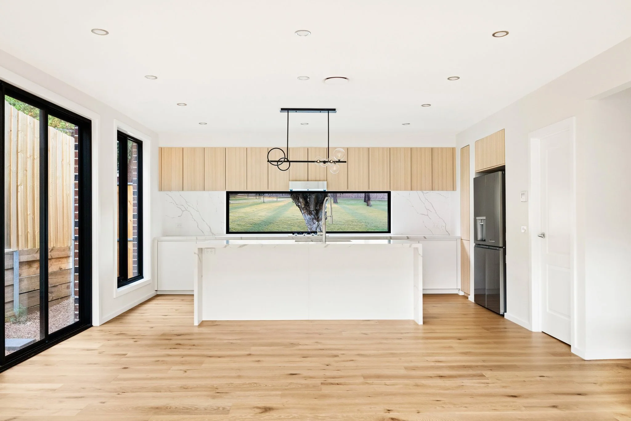 Modern kitchen with wooden cabinets, white marble backsplash, large window showing outdoor grass and trees, black refrigerator, and a central island with a sink, illuminated by recessed ceiling lights.