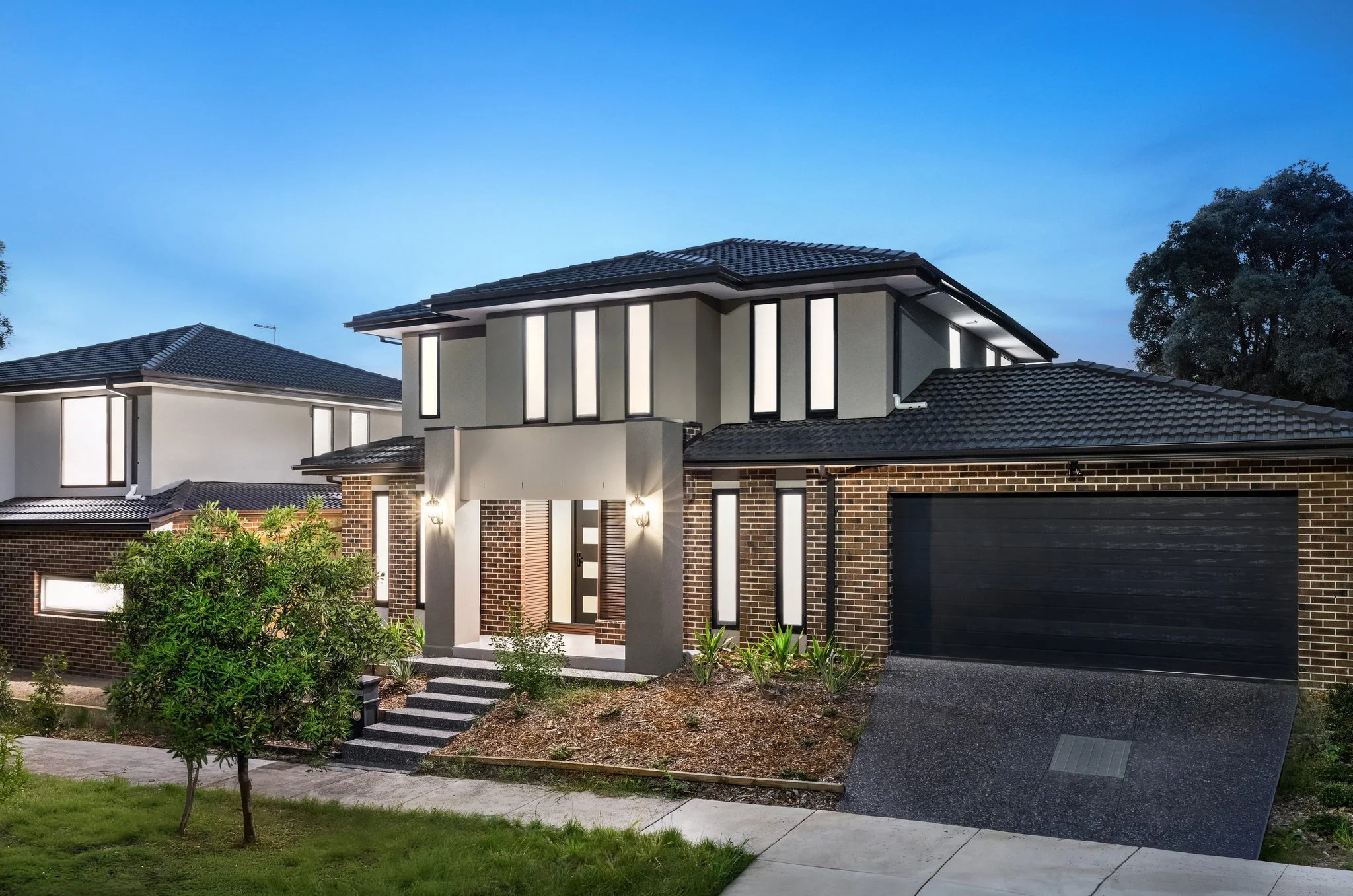 Modern two-story house with brick and grey panel exterior, illuminated windows, black garage door, small front garden with plants and a tree, sidewalk, and a blue sky evening background.