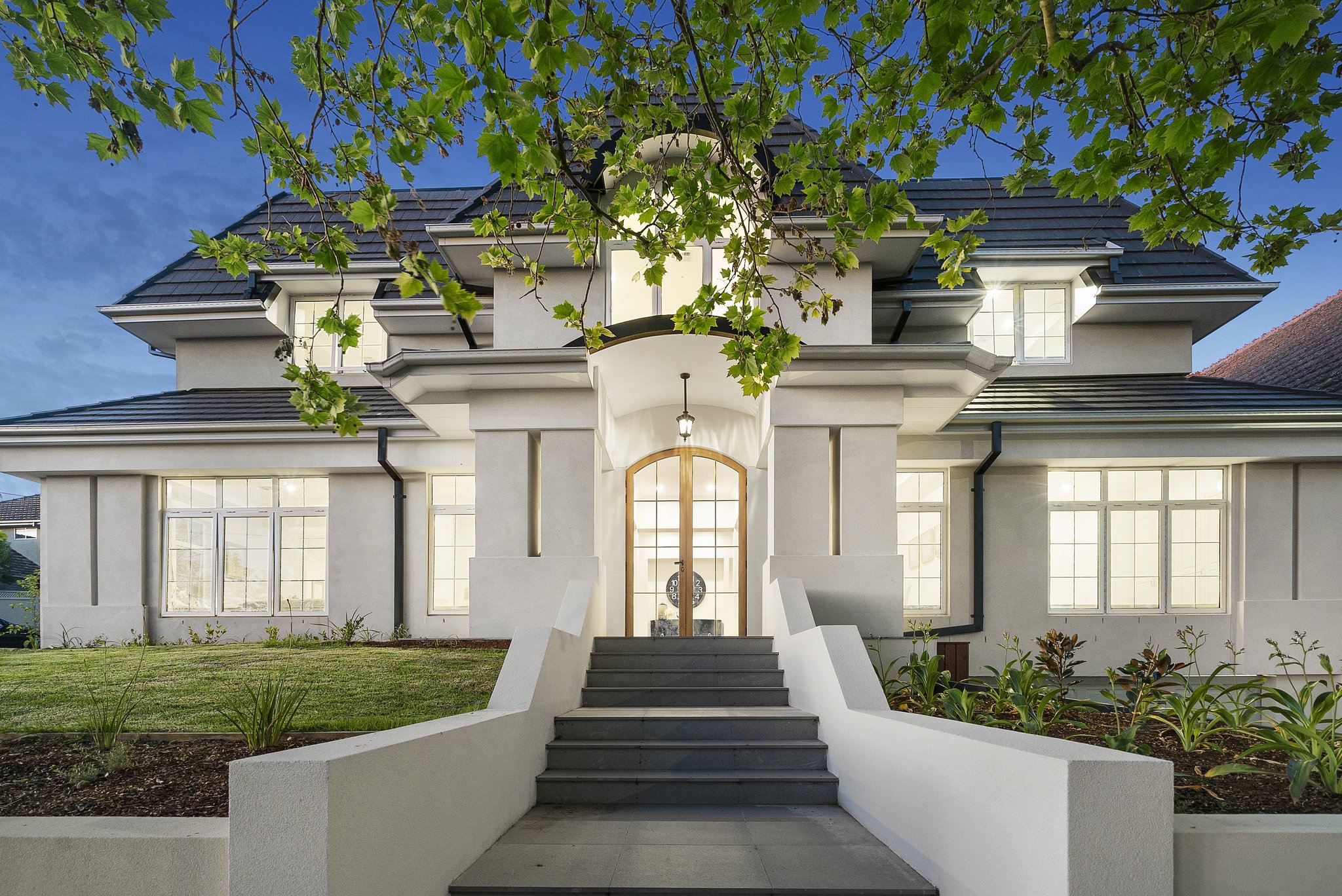 Exterior view of a large, elegant house with a white facade, multiple windows, and a central staircase leading up to the front door, surrounded by landscaped garden beds and green grass, under a blue evening sky with tree branches in the foreground.