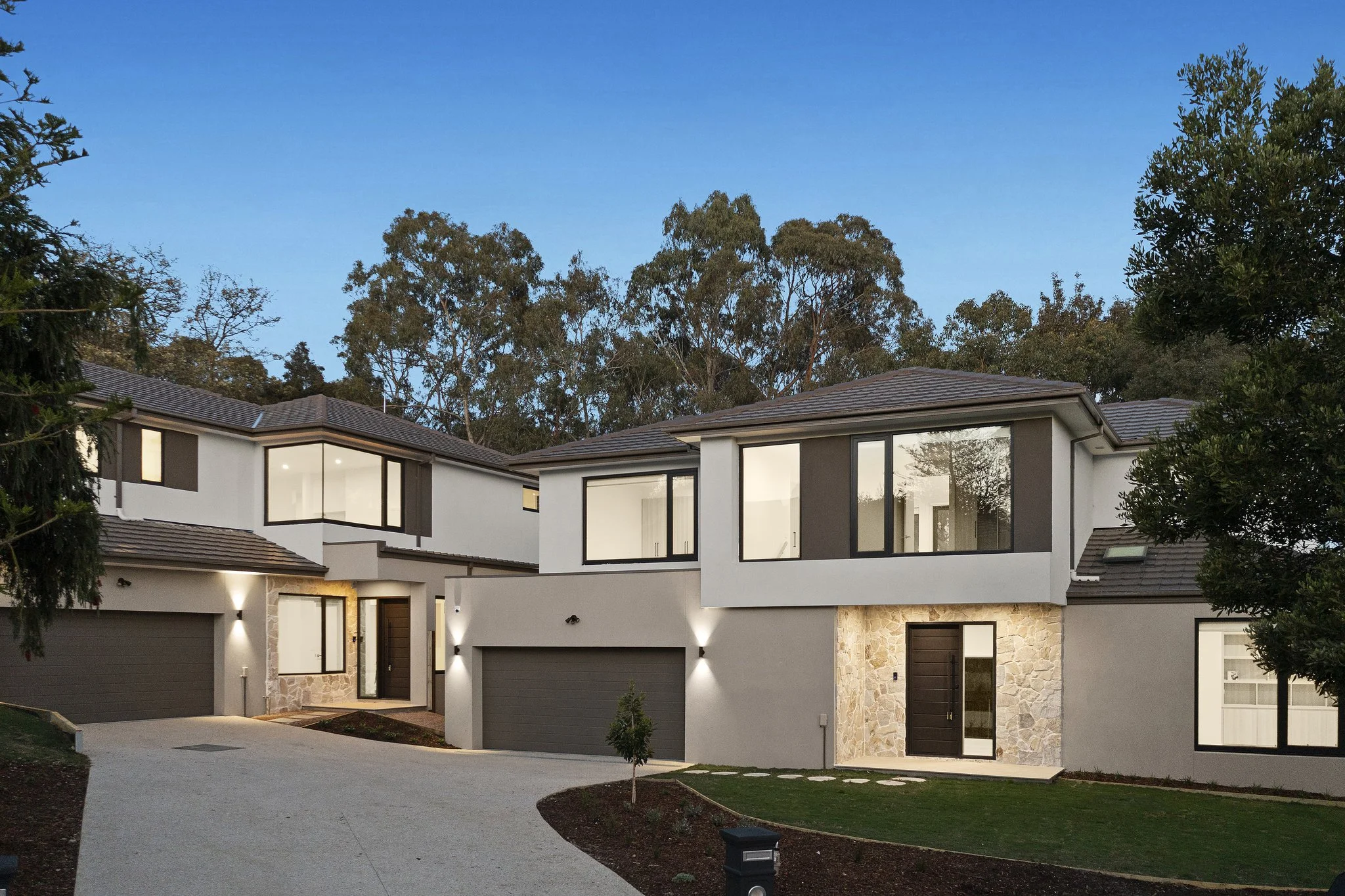 Modern multi-story house with large windows, stone and stucco exterior, surrounded by trees and a curved driveway at dusk.