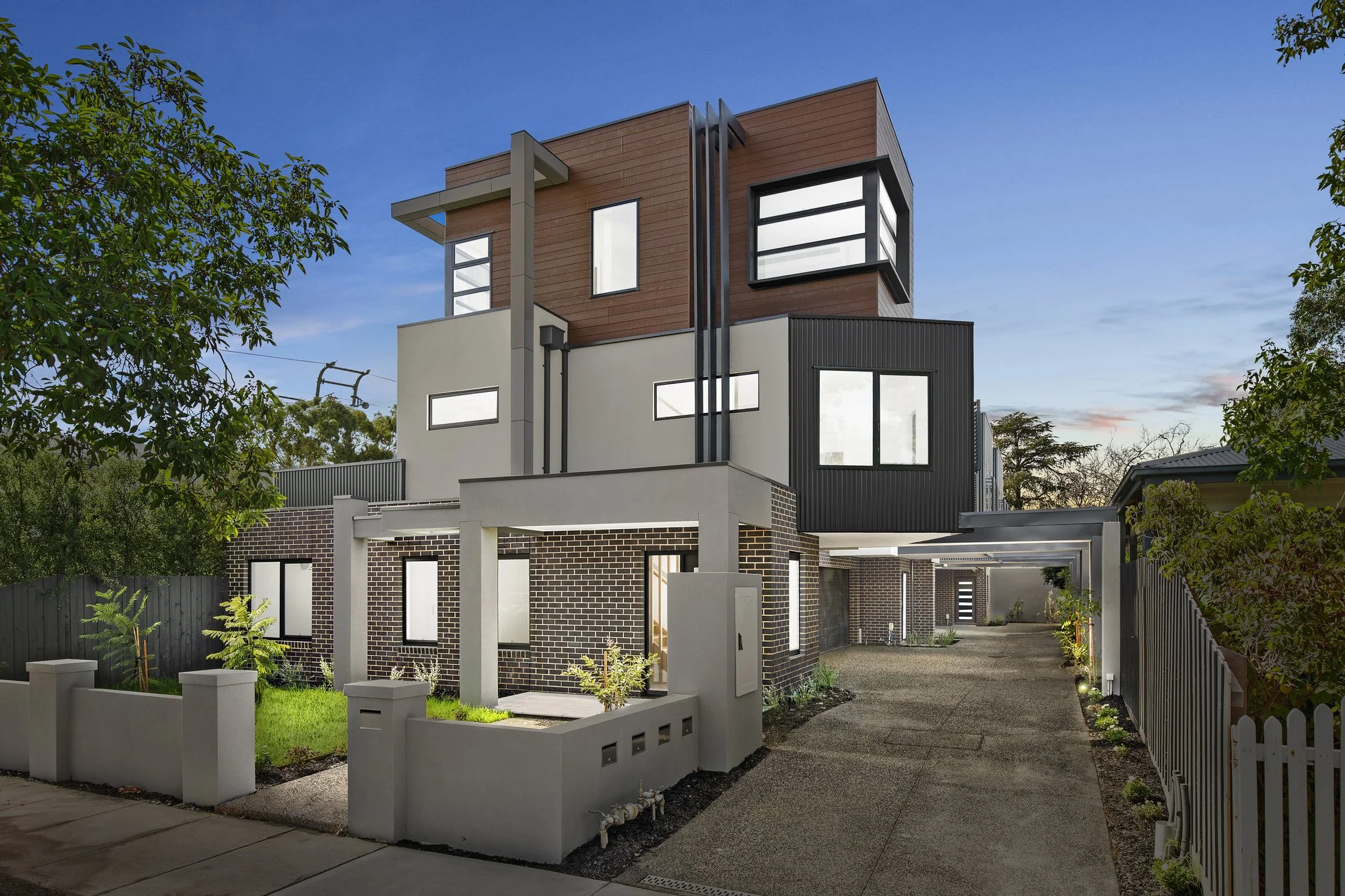 Modern multi-story house with brick, gray, black, and wood exterior finishes, surrounded by a small yard with plants, and a driveway on the right, under a clear blue sky.