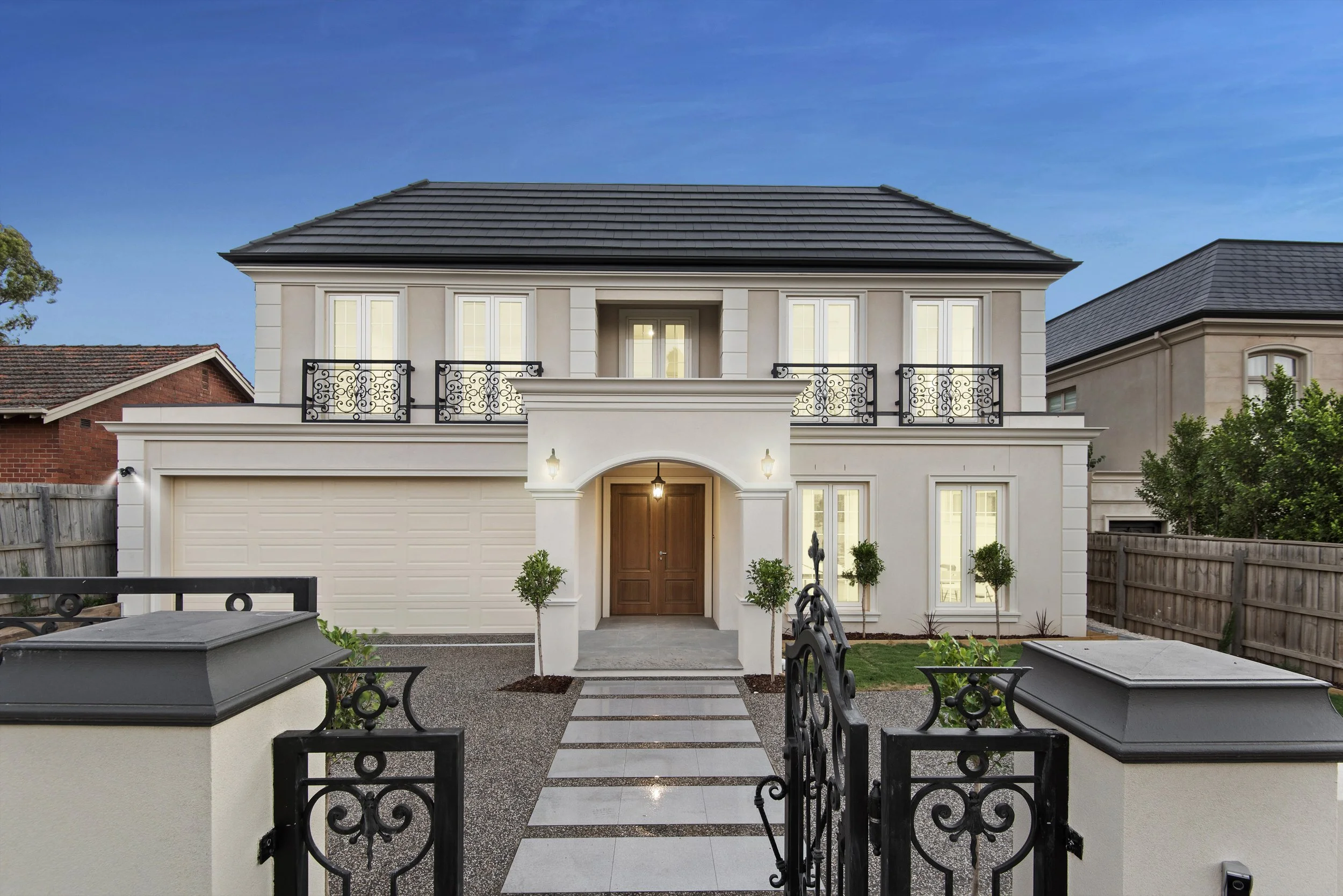 Front view of a modern, two-story house with a white exterior, black wrought iron balcony railings, a brown wooden front door, and a gated entry with a paved walkway and small trees.