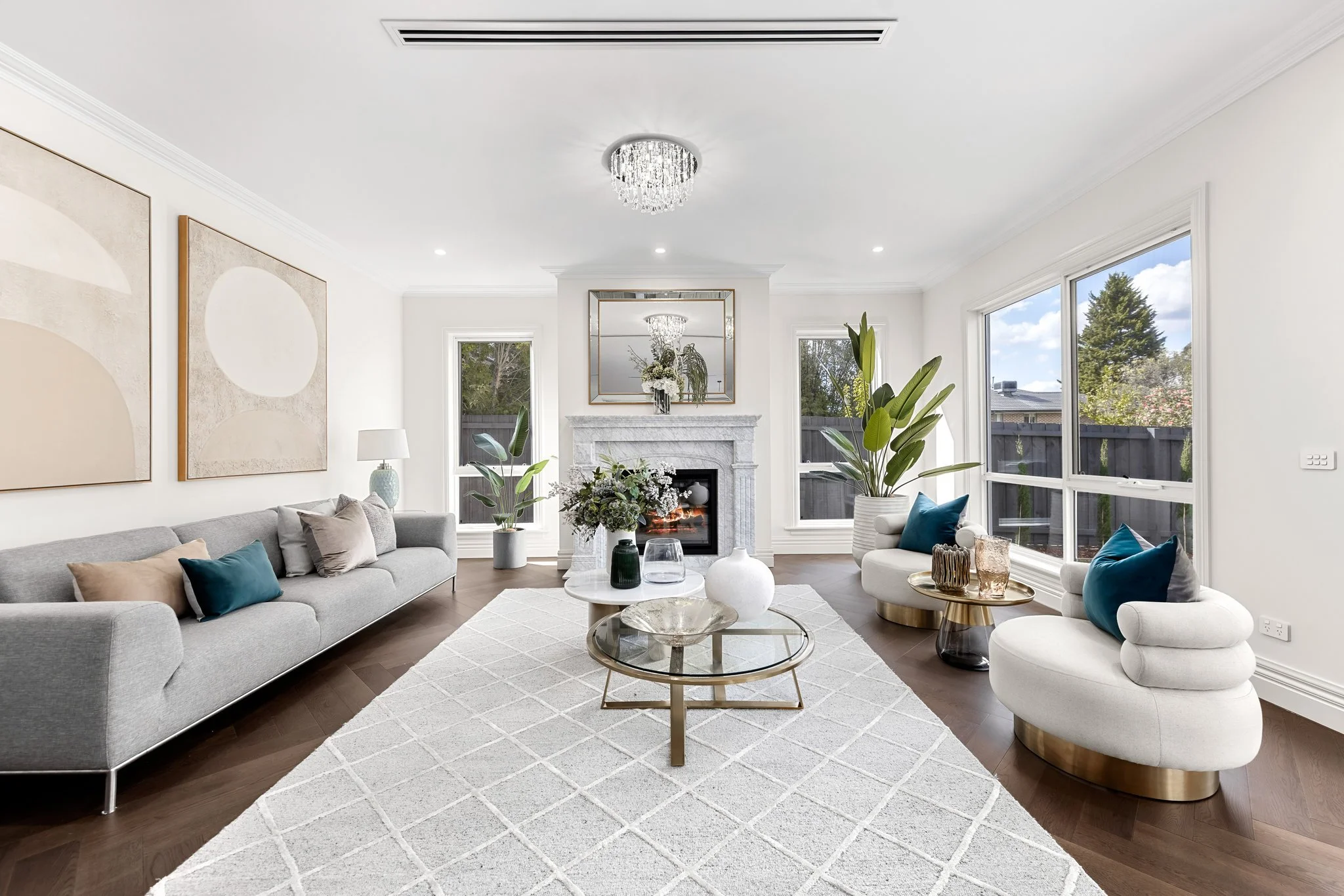 Bright living room with white walls, large windows, a stone fireplace, and contemporary furniture including a gray sofa, white and blue armchairs, and glass coffee tables, decorated with potted plants and vases.
