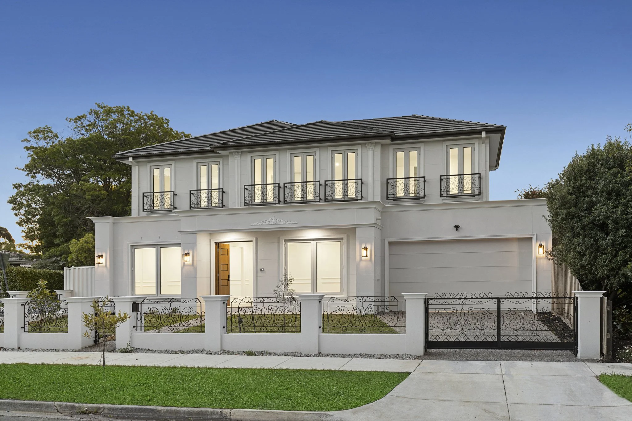 Modern two-story house with white exterior walls, large windows, and black ornate balcony railings, surrounded by a white fence and a green lawn.