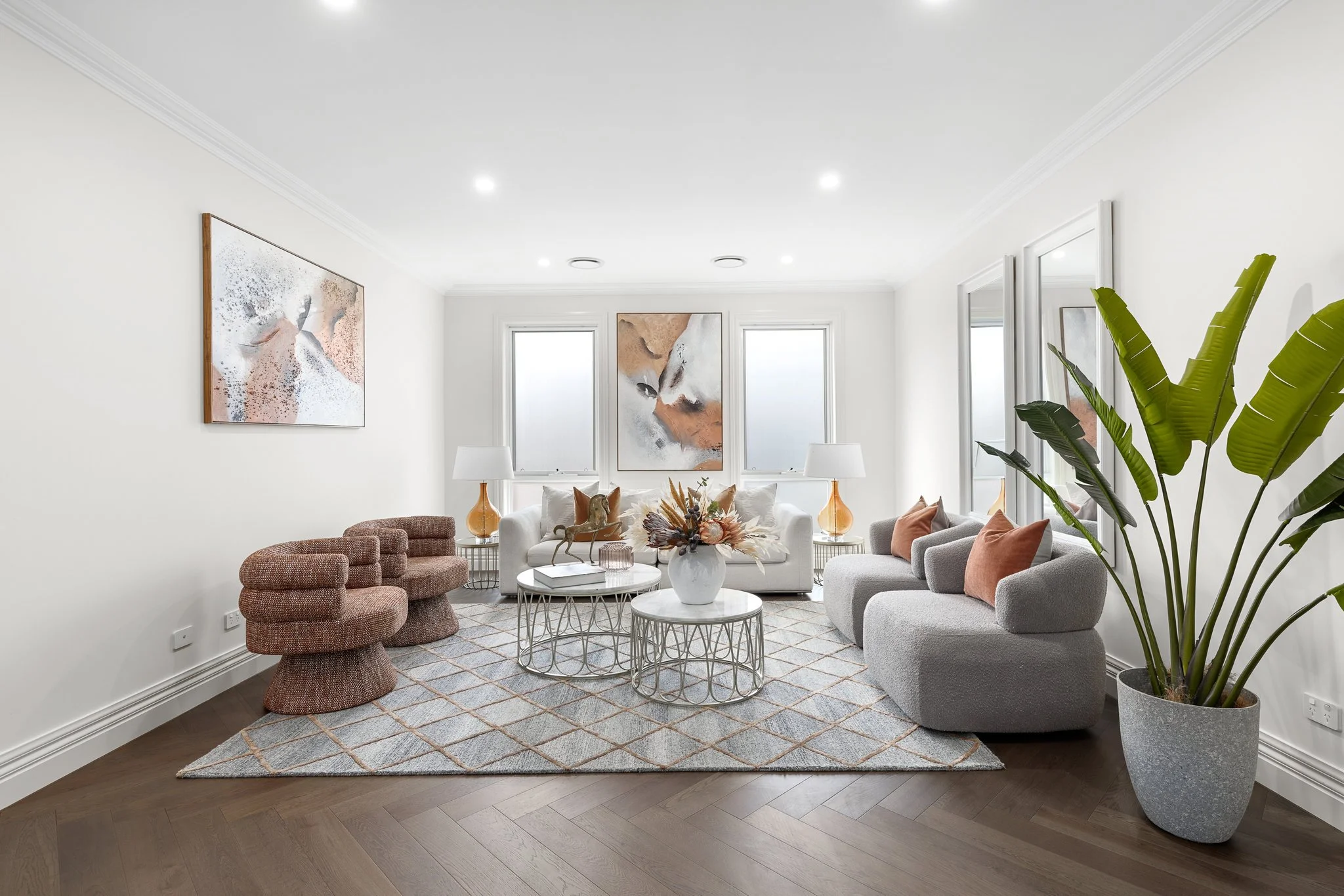 Bright living room with white walls, large windows, and modern furniture. Contains a white couch, two gray armchairs with peach pillows, a textured pink chair, and a large potted plant. Decor includes abstract art on walls, white lamps, a vase with flowers on a central coffee table, and a patterned area rug.