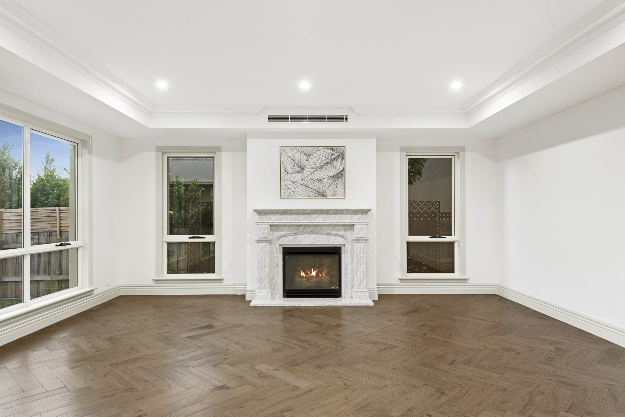 Empty living room with white walls, hardwood floor, a fireplace with a painting above it, large windows on both sides, and ceiling lights.