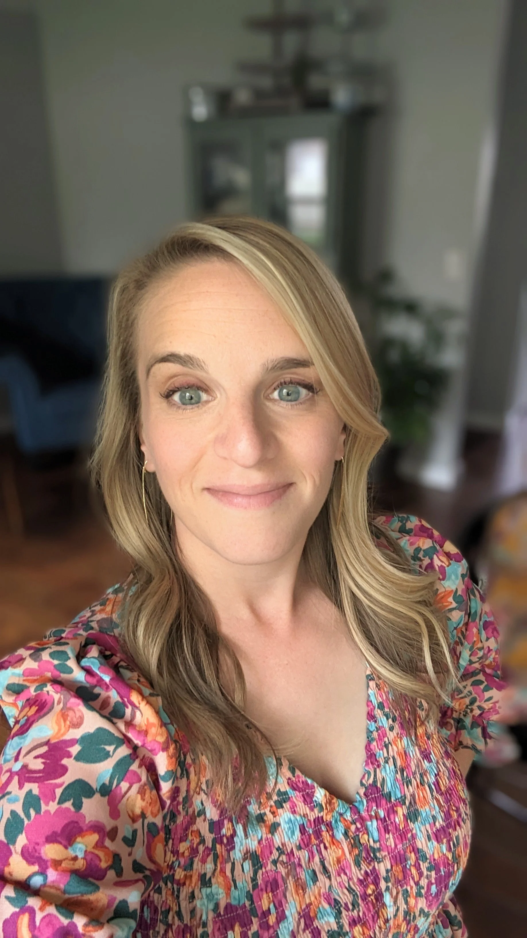 A woman with blonde hair and blue eyes taking a selfie in a room with a blurred background including a blue armchair, a green cabinet, and a potted plant.
