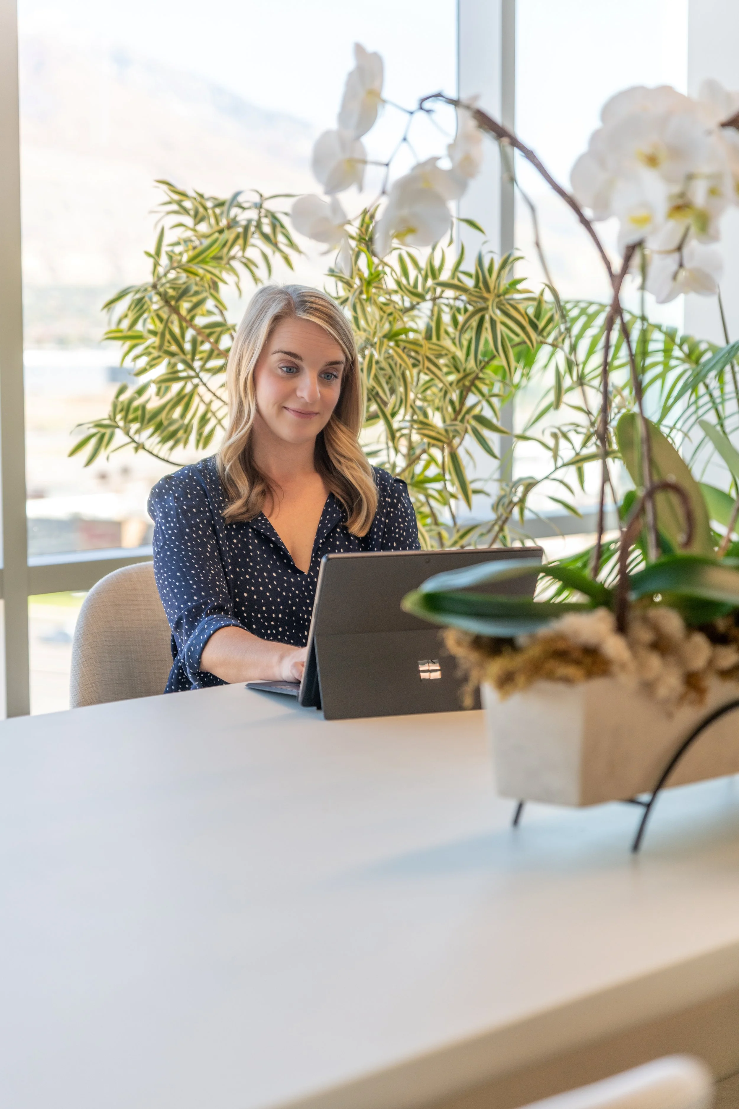 A woman using a Microsoft Surface tablet at a desk with a large houseplant and orchids in an office with large windows.