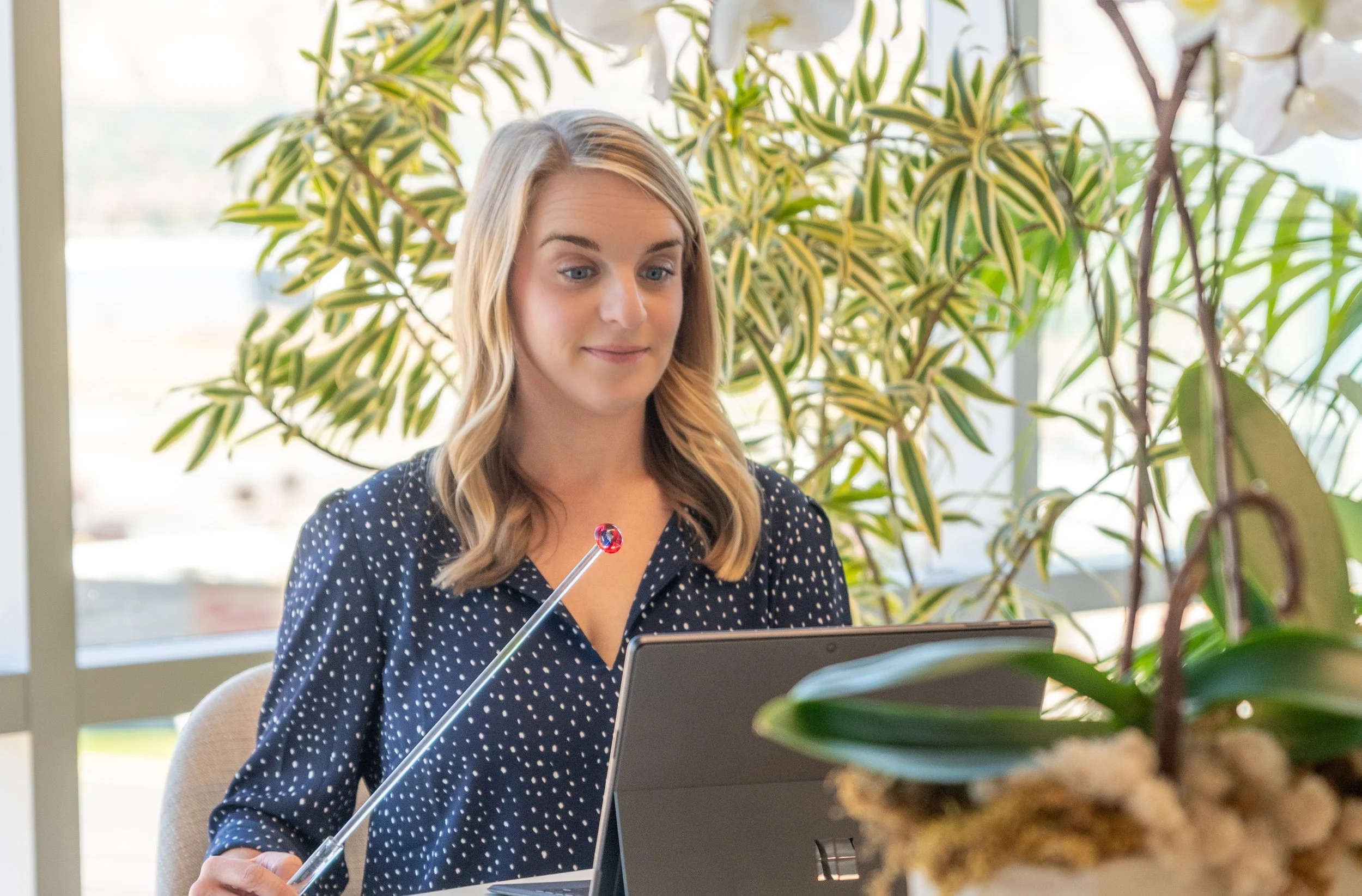 Woman with blonde hair in a navy blue and white polka dot blouse sitting at a desk with a computer, holding a eye directing device, surrounded by potted plants.