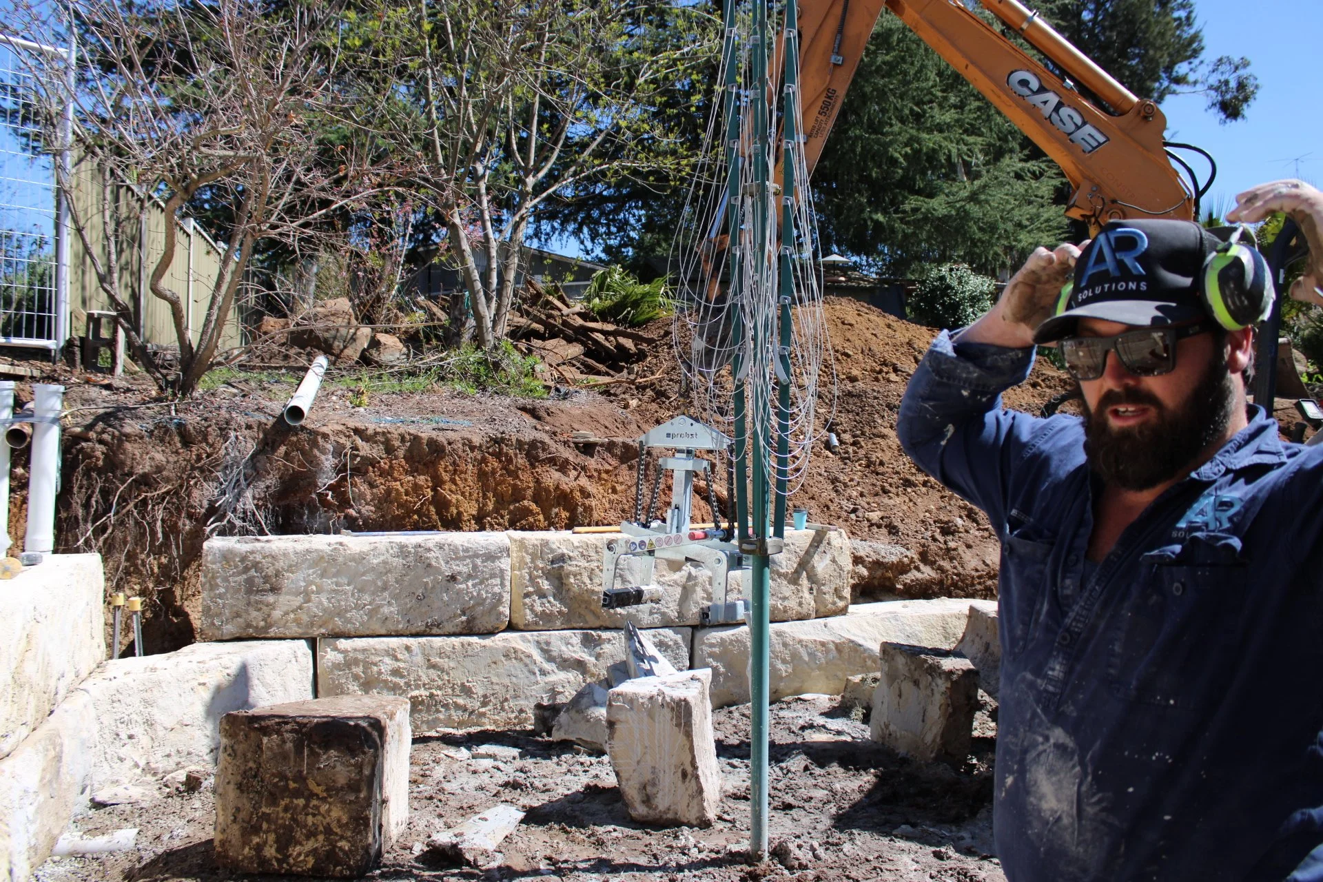 A construction worker wearing safety glasses, a helmet, and noise-canceling headphones is standing on a construction site, looking to the side. Behind him, there is a pile of dirt, some stone blocks, an excavator, and trees in the background.