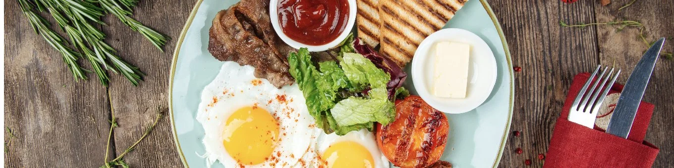 Breakfast plate with eggs, toast, grilled tomato, bacon, salad, butter, and ketchup on a wooden table.