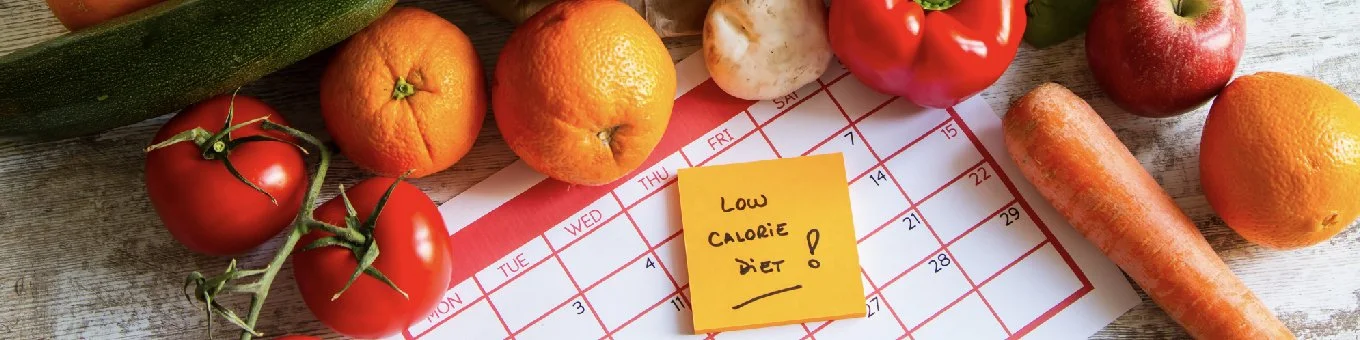 Fresh fruits and vegetables, including tomatoes, oranges, a zucchini, a carrot, and an apple, placed on a wooden surface near a calendar page with a yellow note that says 'Low Calorie Diet!'