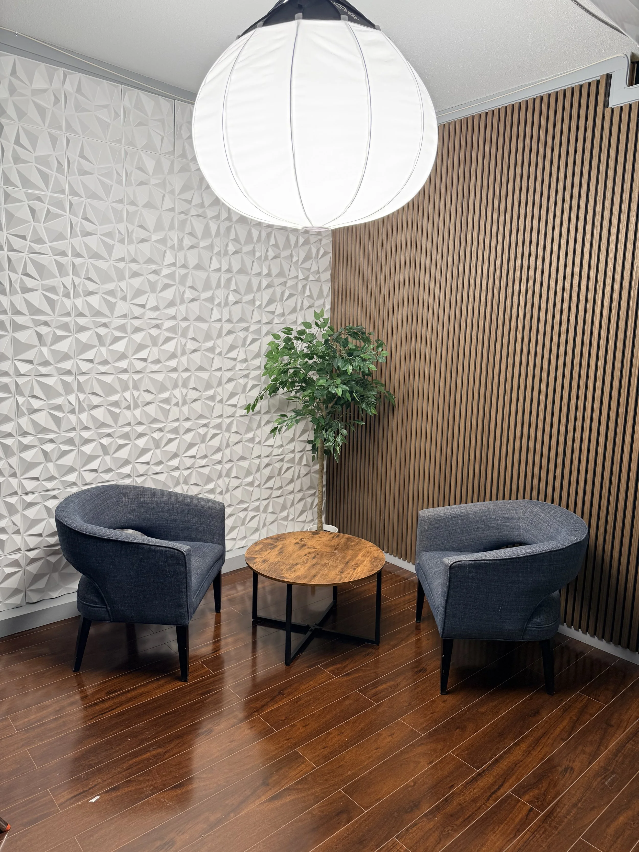 Two dark gray upholstered armchairs surrounding a small round wooden table, with a potted green plant behind them, in a modern room with textured white and wooden wall panels and a large white pendant light.