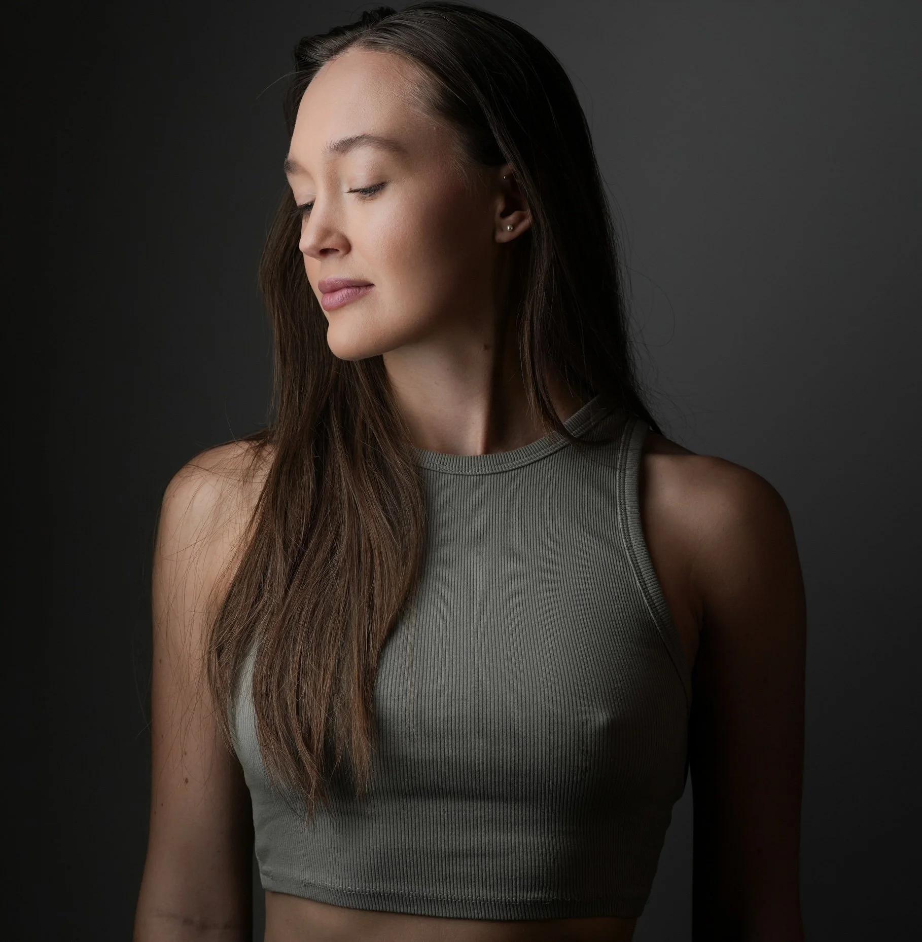 Young woman with closed eyes, long brown hair, wearing a sleeveless gray top, standing against a dark background.