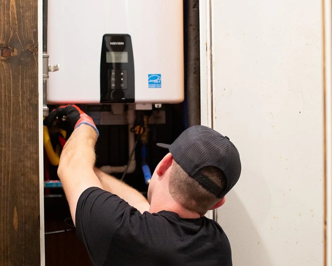 A man working on a water heater or boiler, wearing a black cap and gloves, in a small utility closet.