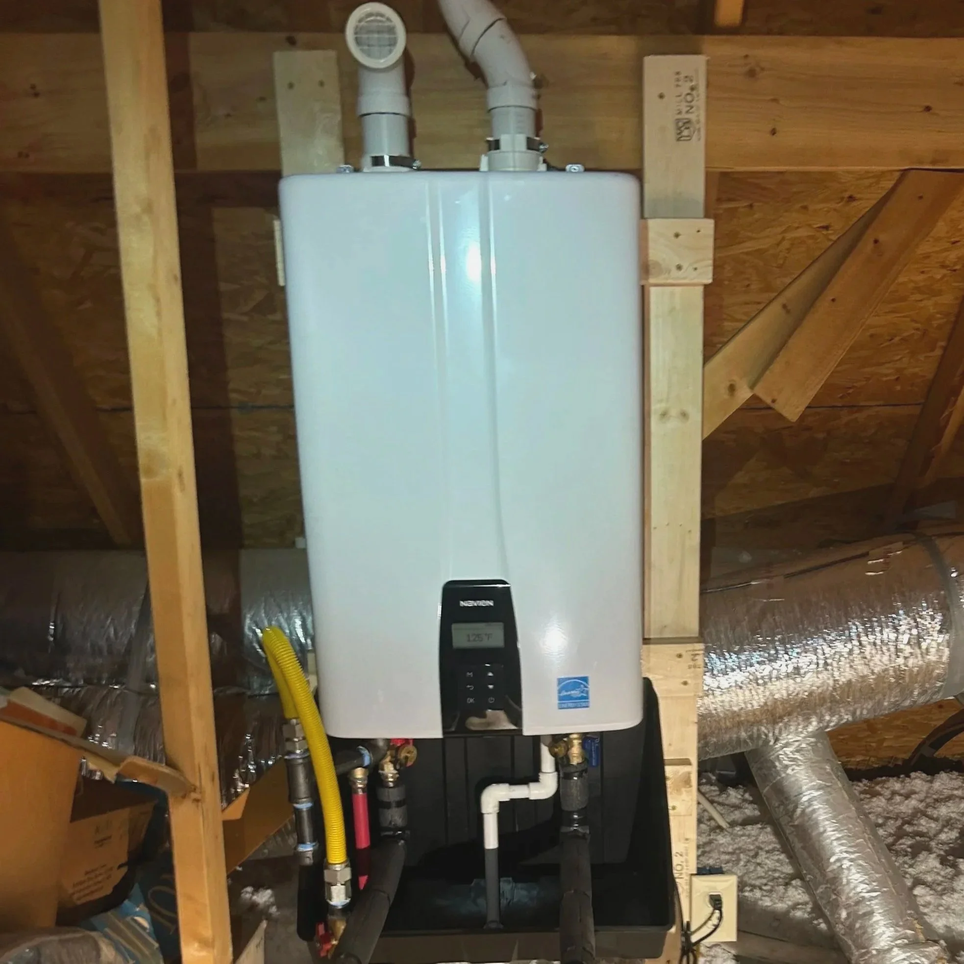 A white tankless water heater mounted on an unfinished wall in an attic space, with pipes and ductwork connected below and around it.