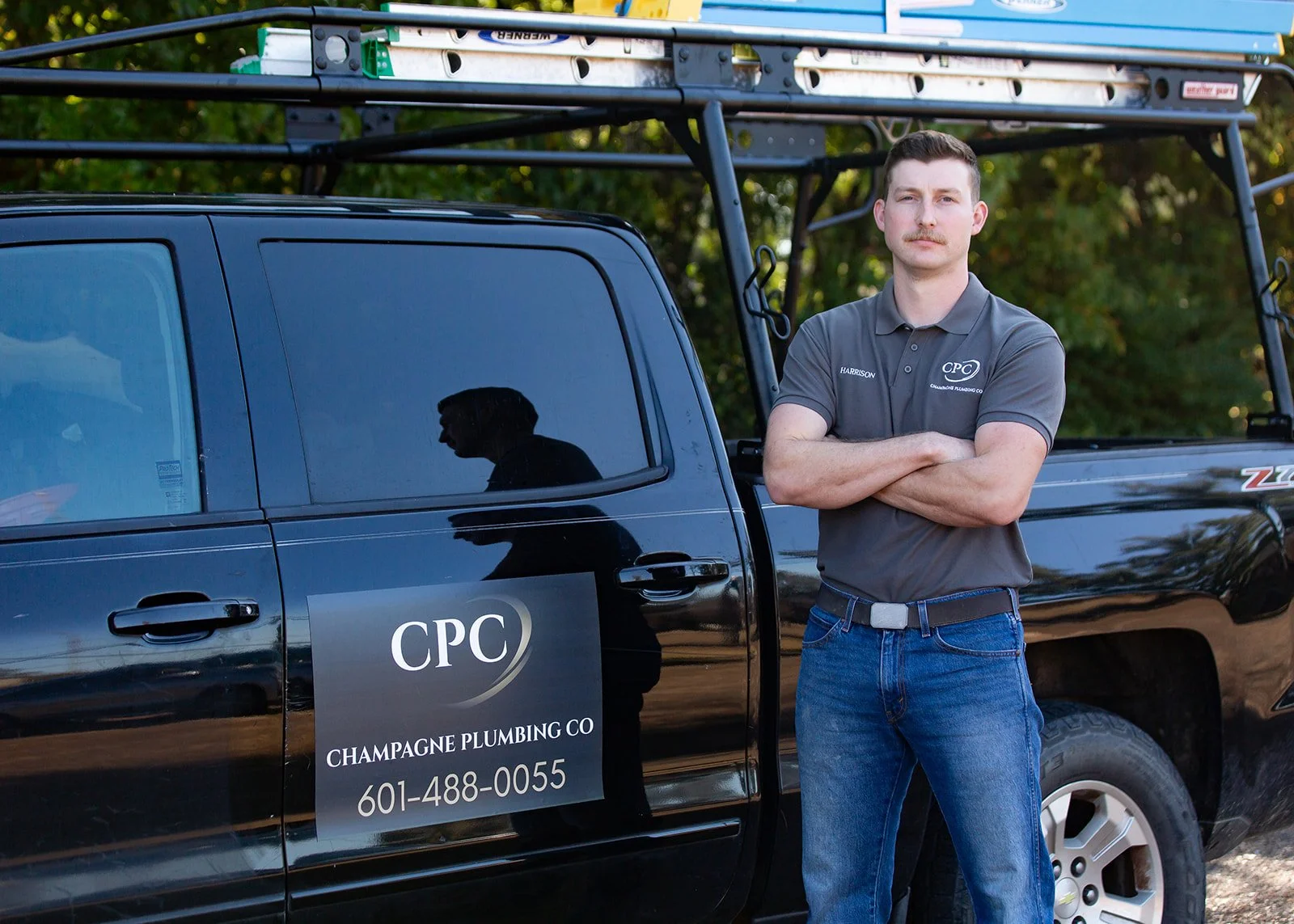 A man standing with arms crossed in front of a black pickup truck marked with the logo and contact information of Champagne Plumbing Co.