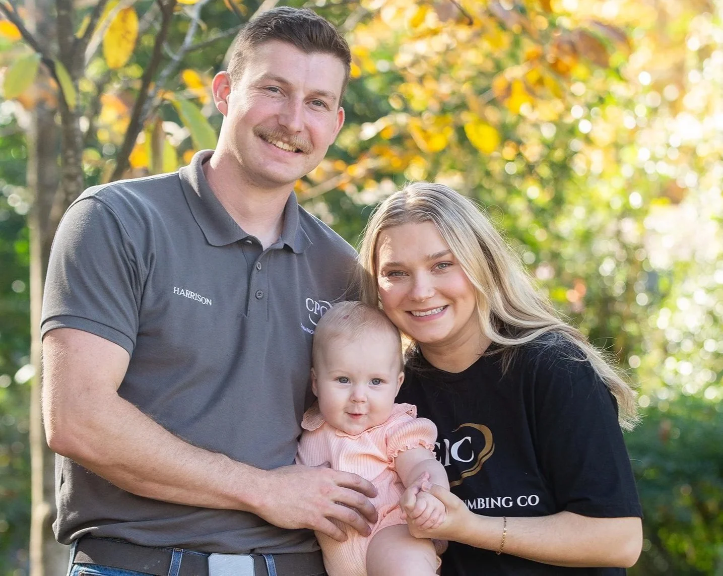 A smiling family consisting of a man, woman, and baby outdoors during daytime, with trees and sunlight in the background.