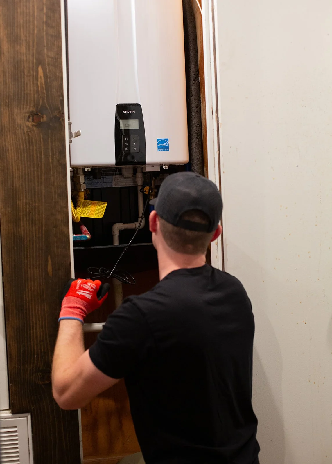 A man wearing a black shirt, a grey baseball cap, and red gloves working on a water heater or boiler inside a closet or cabinet.