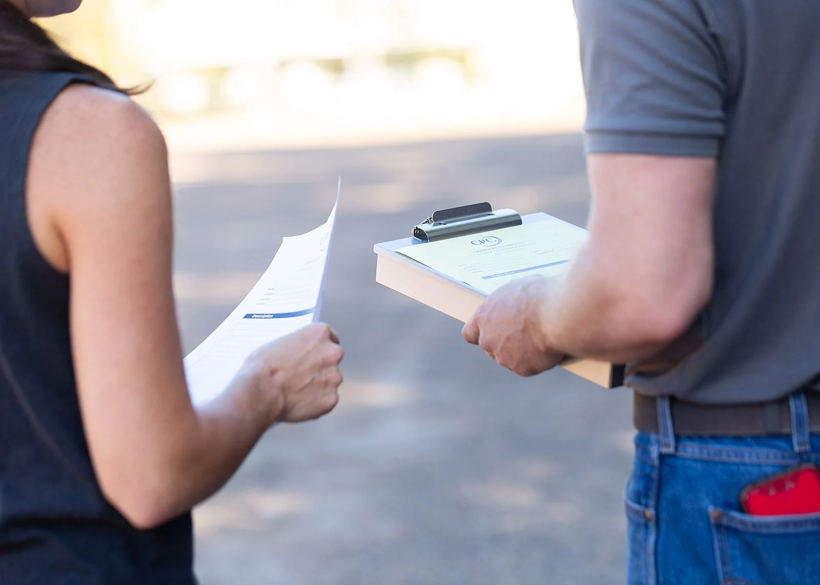 Two people standing outdoors exchanging papers and a clipboard, one holding a document, the other holding a clipboard with a form on it.