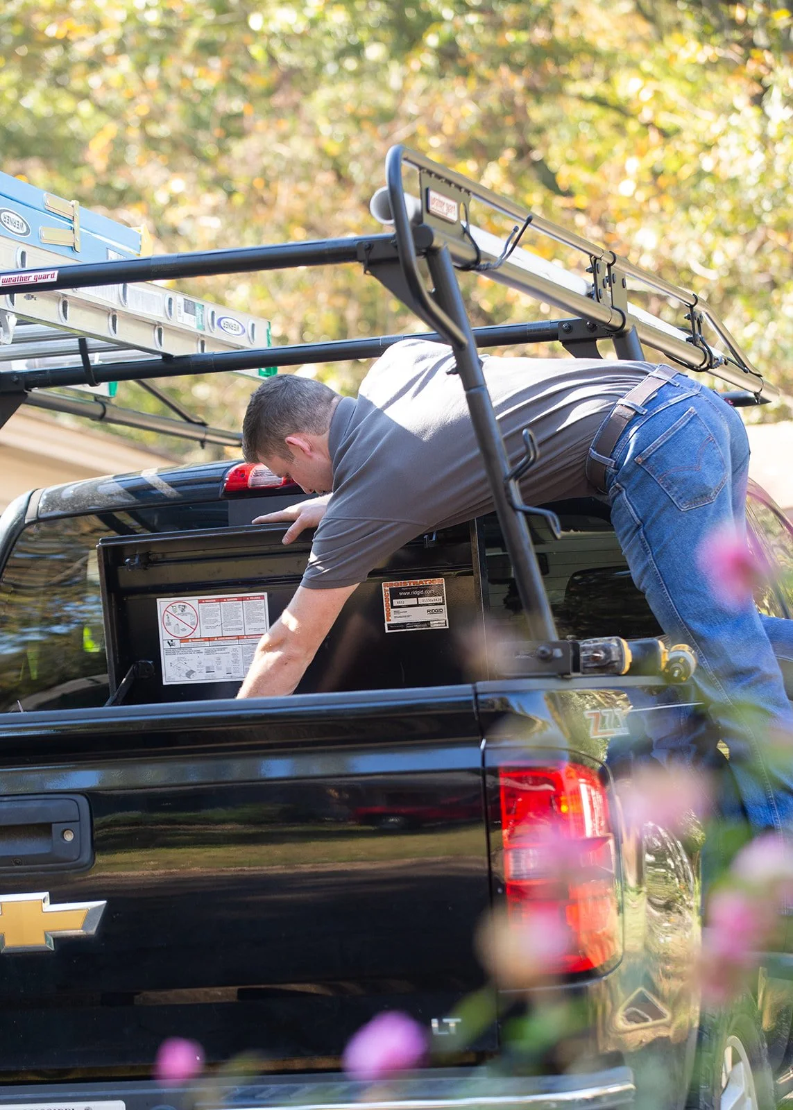 A man leaning into the open tailgate of a black Chevrolet pickup truck, with ladder and equipment on the roof, in a setting with trees and colorful foliage.