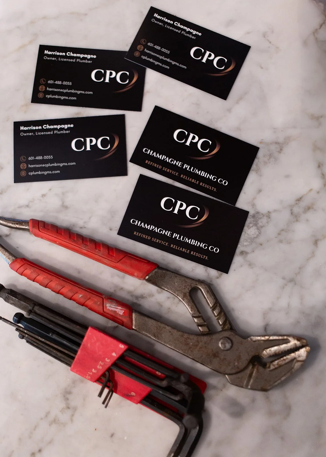 Business cards for Champagne Plumbing Co. featuring the name Harrison Champagne, contact information, and website. Plumbing tools including a pipe wrench and hex keys are also on a white marble surface.
