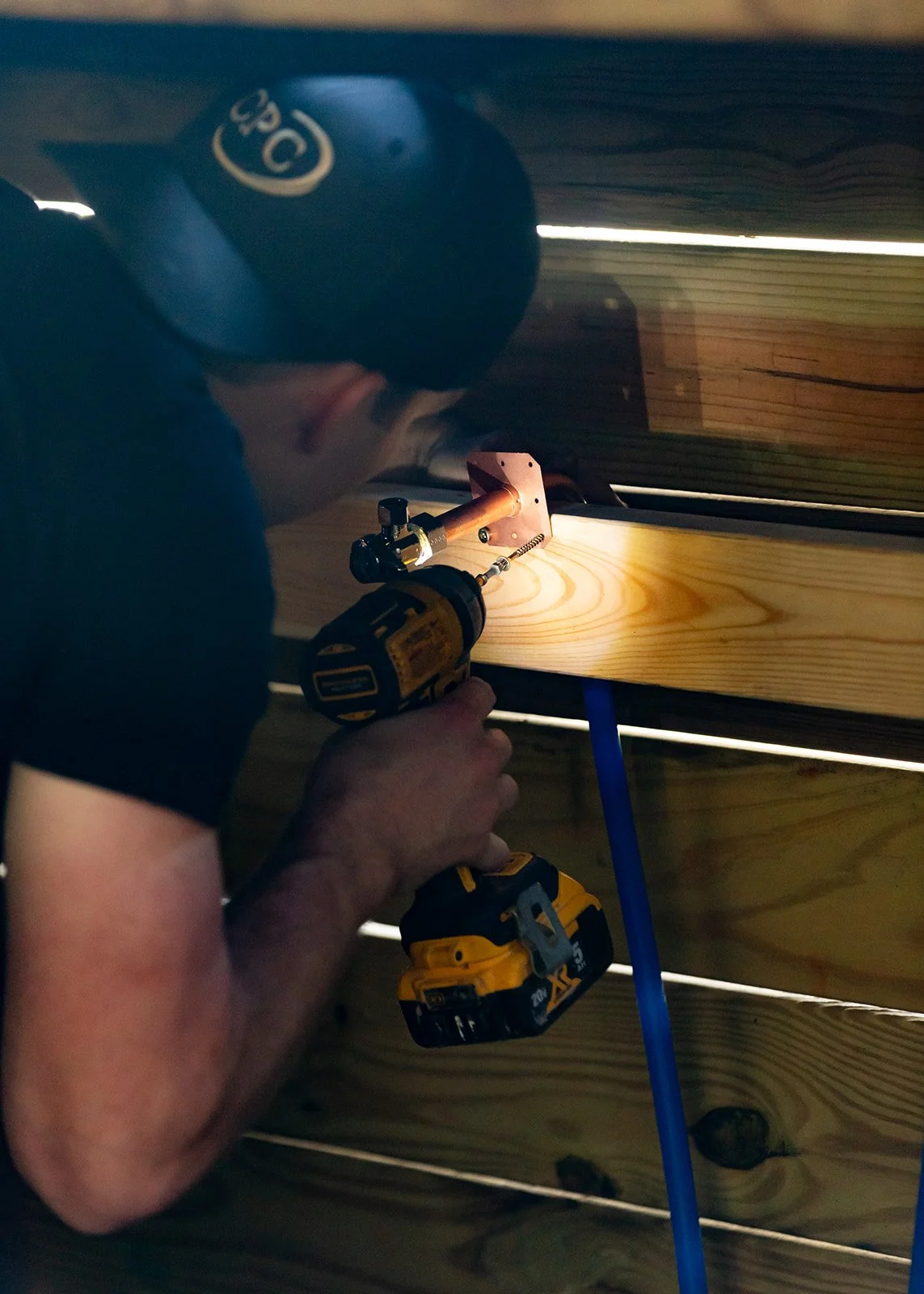 A person wearing a black cap and black shirt is using a cordless drill to secure a copper pipe to a wooden beam inside a wooden structure.