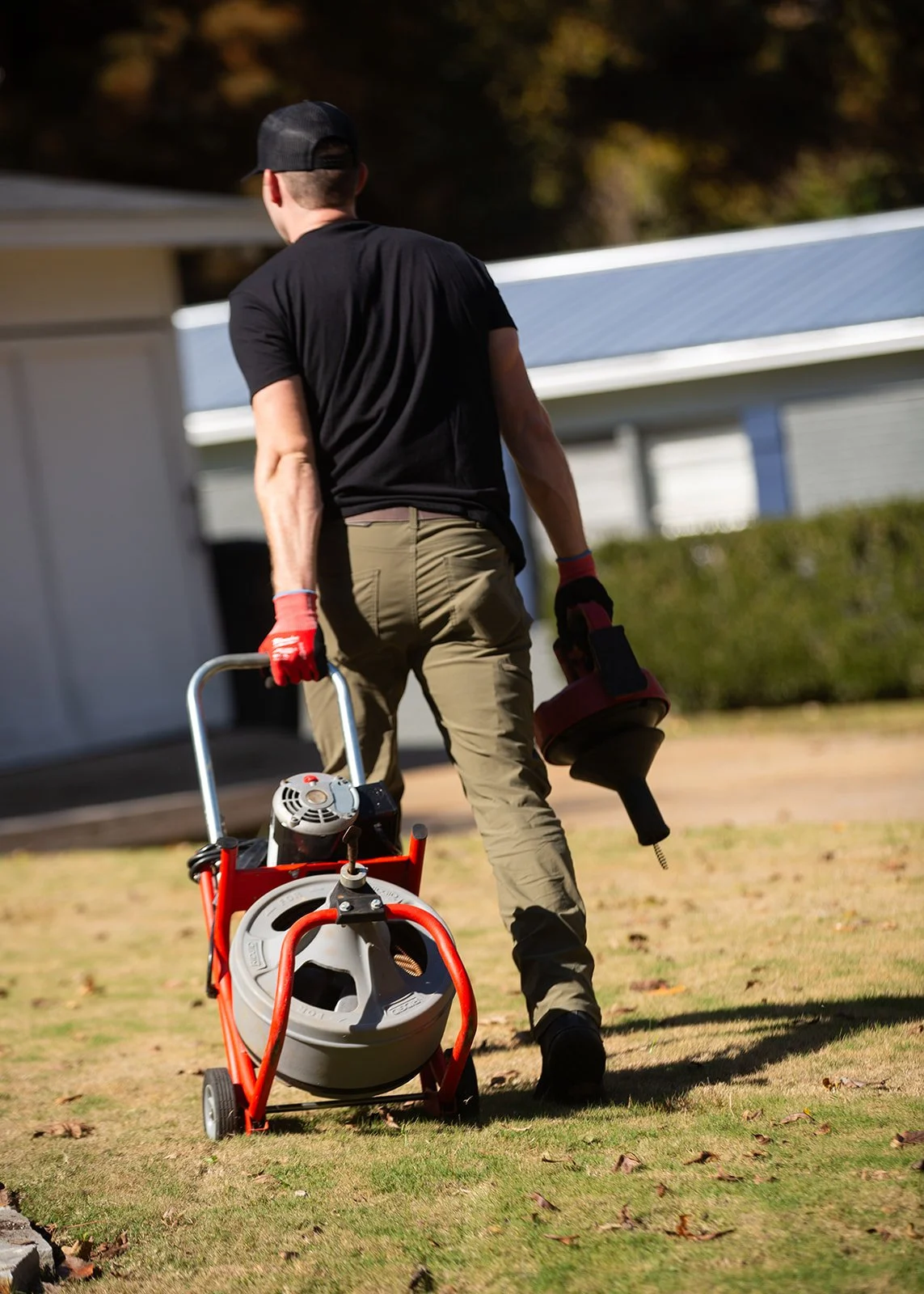 A man wearing a black shirt, khaki pants, and a black cap walks on a lawn pulling a small red and gray lawn trimming machine and carrying a red and black string trimmer.