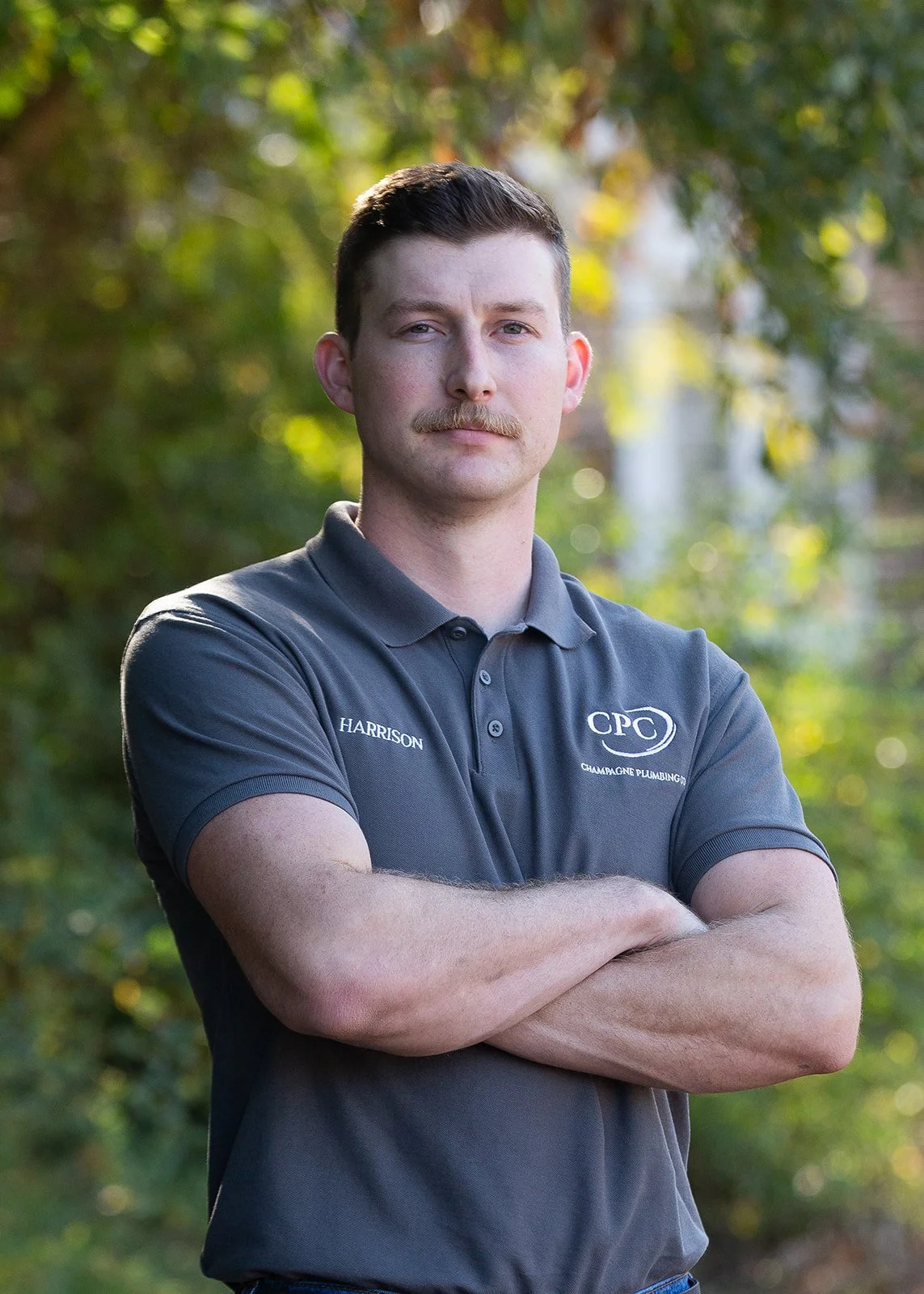 A man with short brown hair, a mustache, and light skin standing outdoors with arms crossed, wearing a dark gray polo shirt with the text 'HARRISON' and a logo on it, in front of a green, blurred natural background.