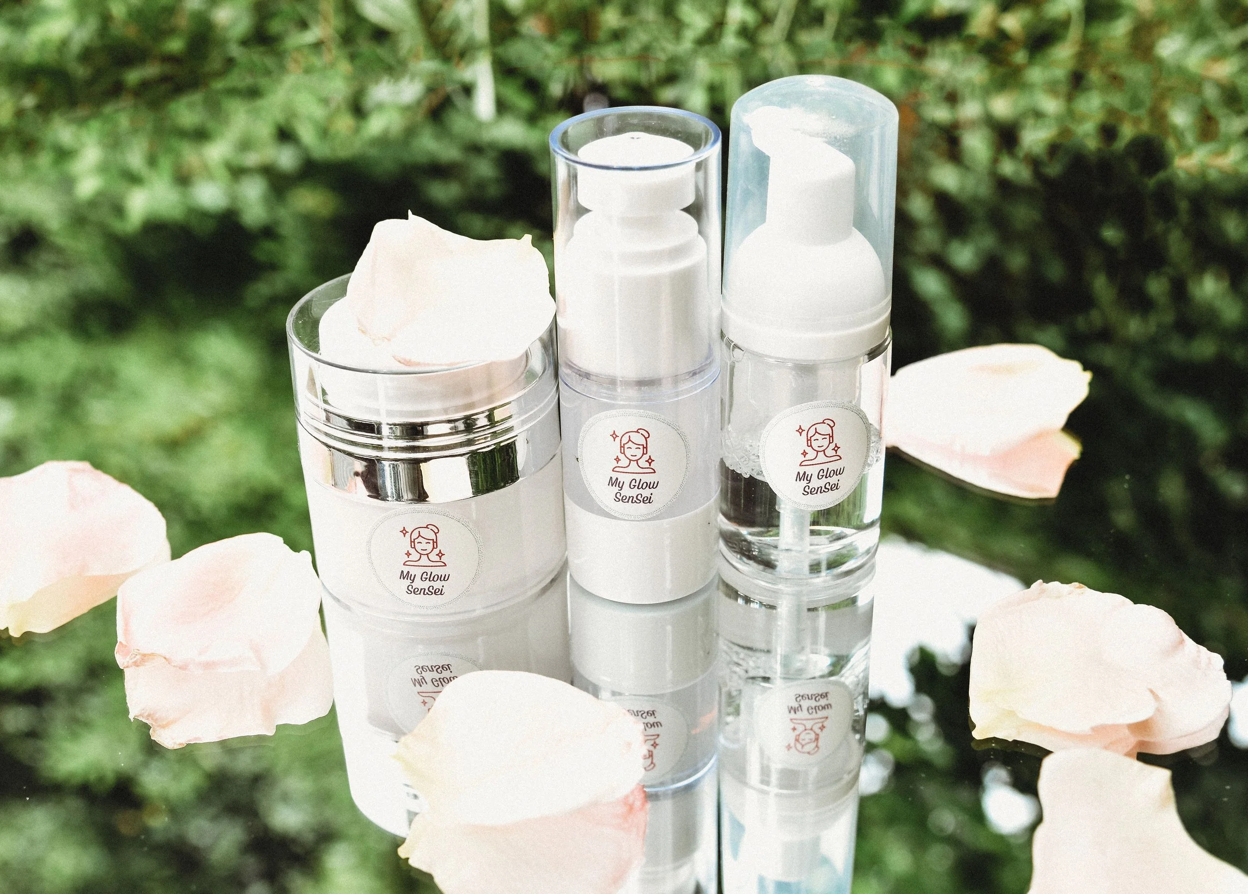 Skincare products in clear bottles and jars labeled 'My Glow Sensei' set on a reflective surface surrounded by pale pink rose petals, with green foliage in the background.