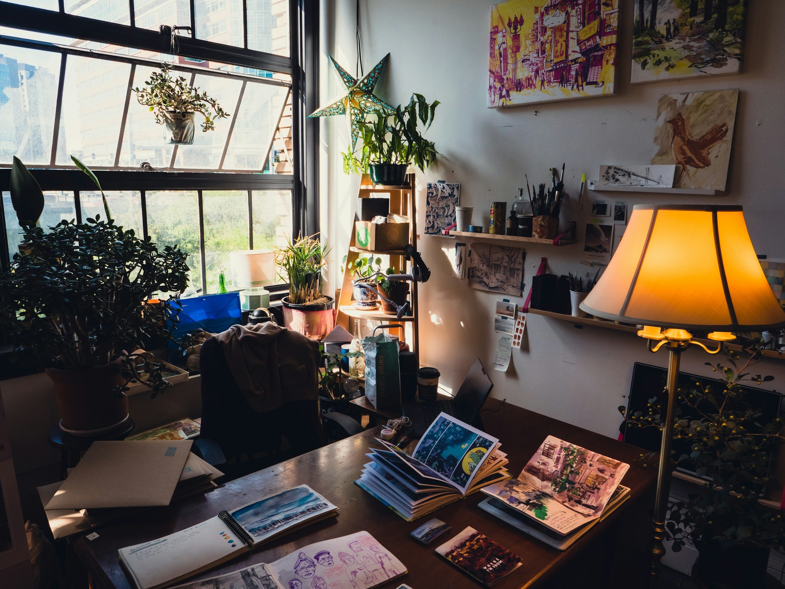 A cluttered art studio with a bright window, potted plants, art supplies, books, and artwork on the walls. A table with open sketchbooks and a tall floor lamp.