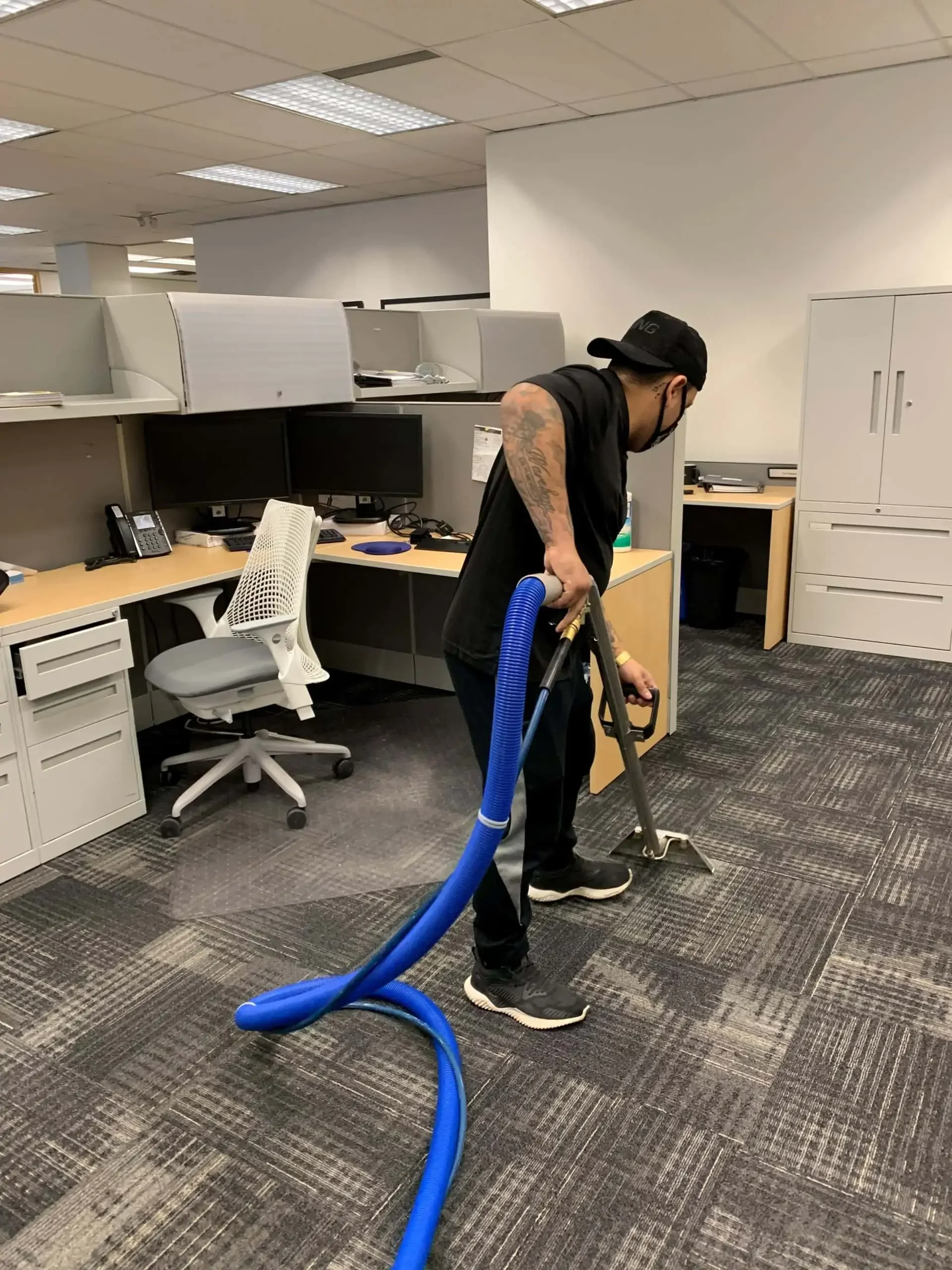 A man vacuuming an office carpet with a blue vacuum hose. The office has cubicles, computer monitors, and office chairs.