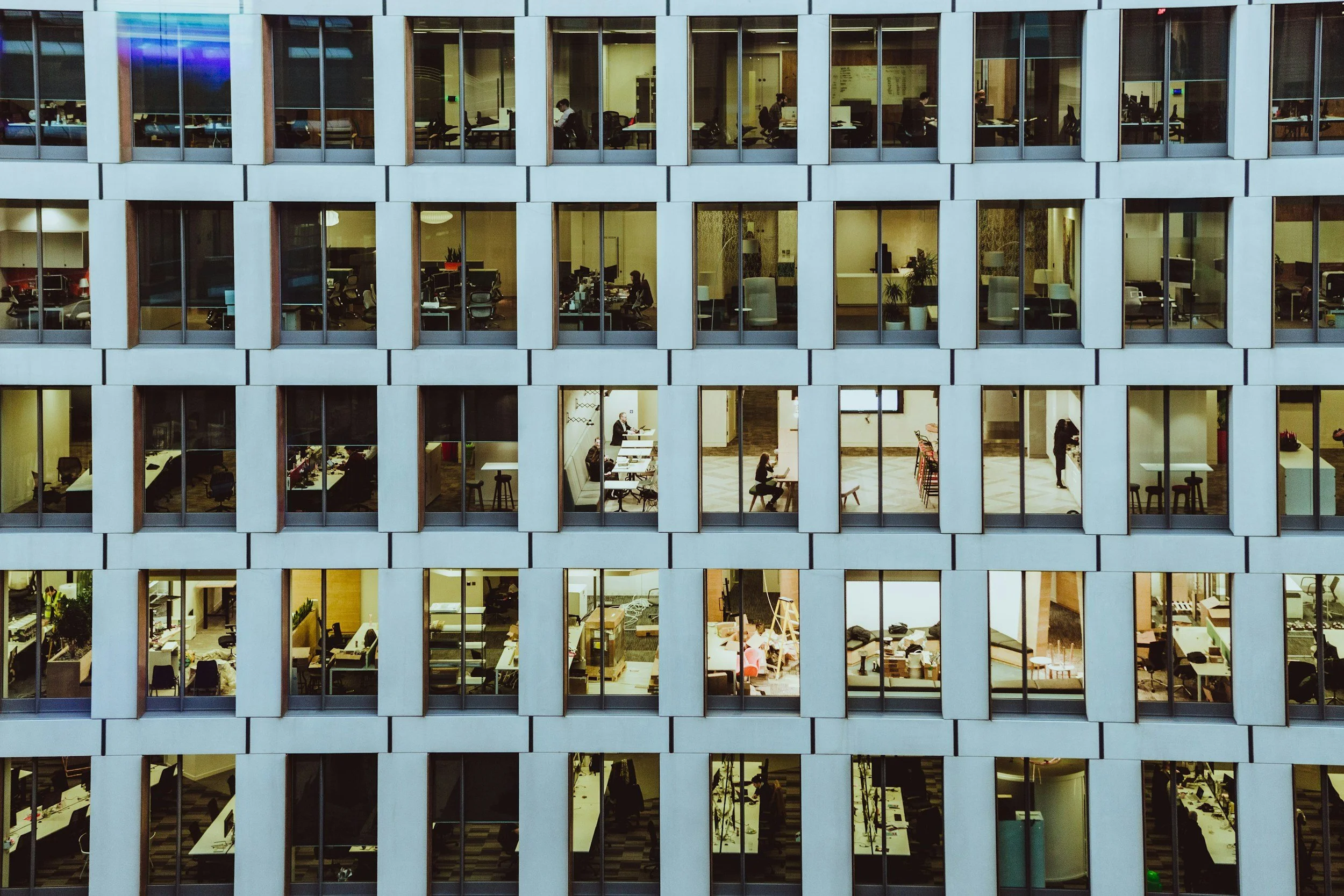 The photo shows the interior of a modern office building from outside with multiple floors visible through large glass windows, with workers working at desks and meeting areas.