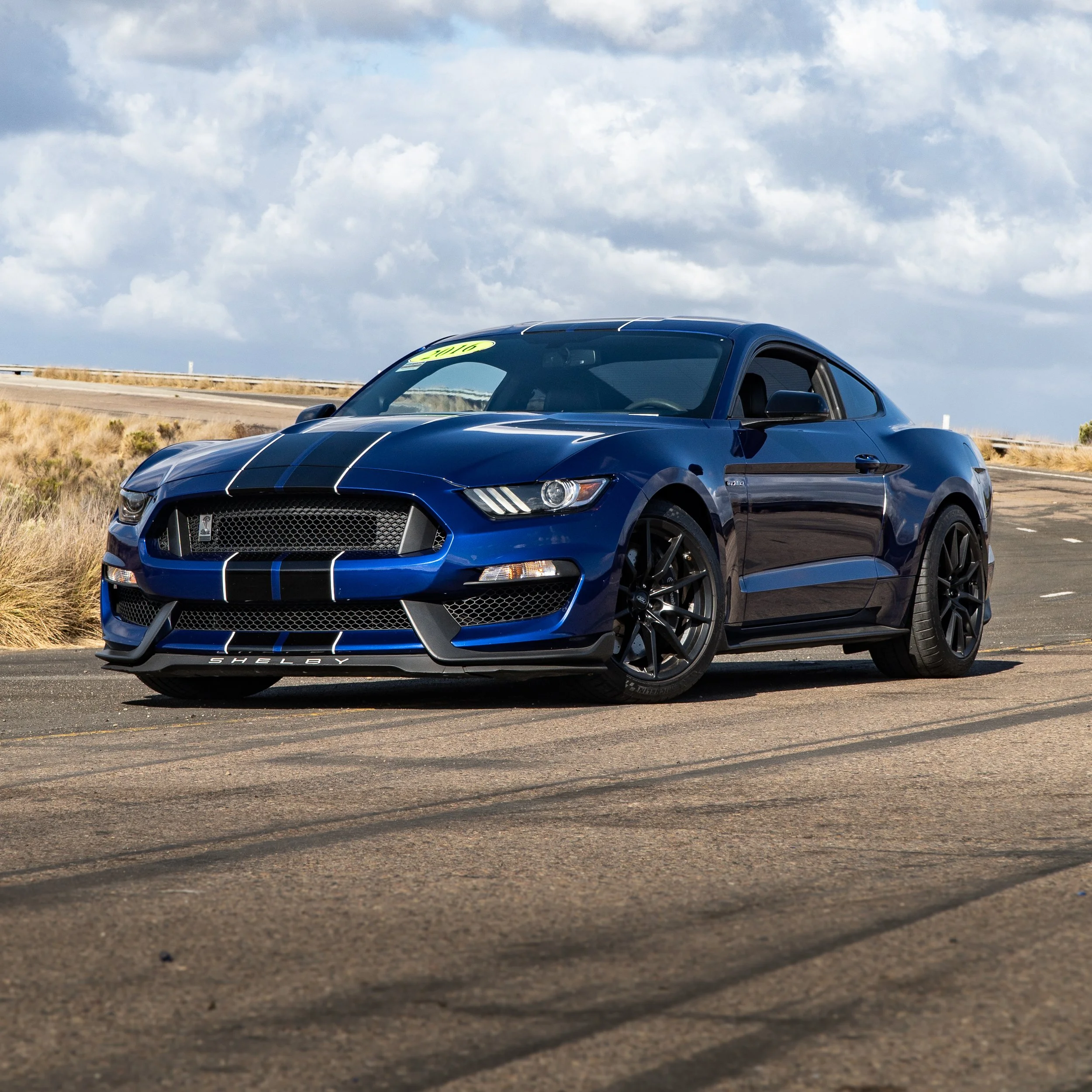 Blue Ford Mustang sports car parked on an open road with a cloudy sky in the background.