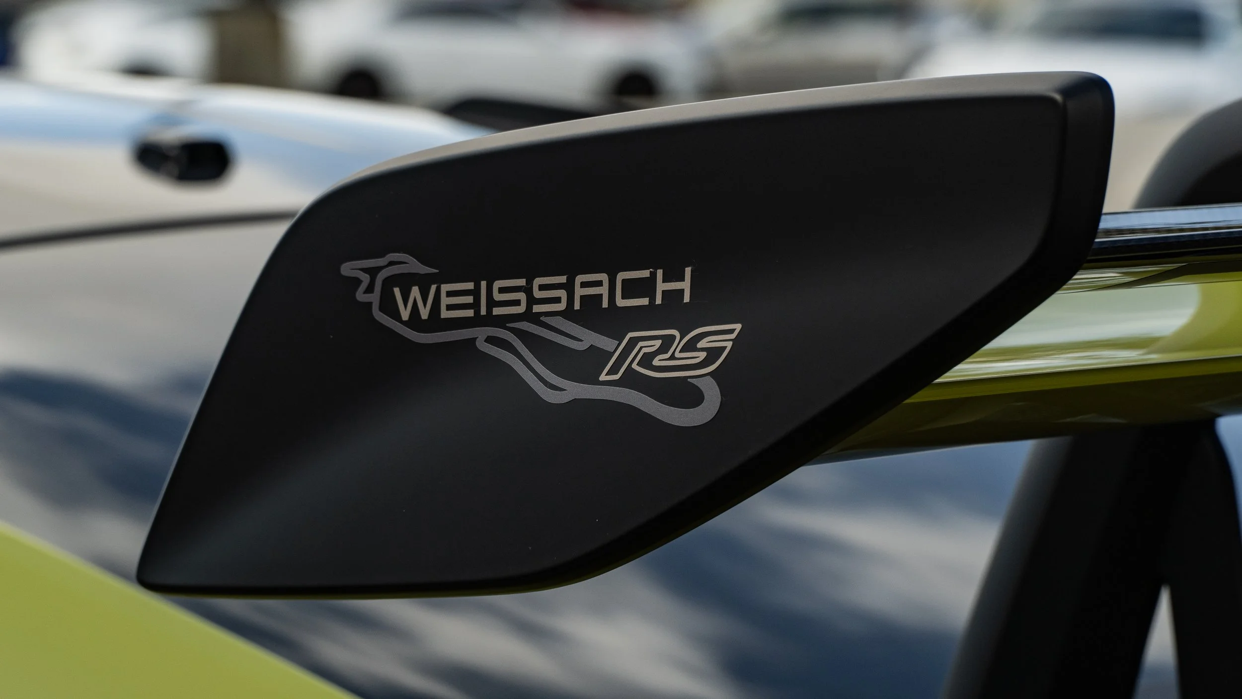 Close-up of a black rear wing with the 'Weissach RS' logo on it, attached to a high-performance sports car. The background shows other cars.