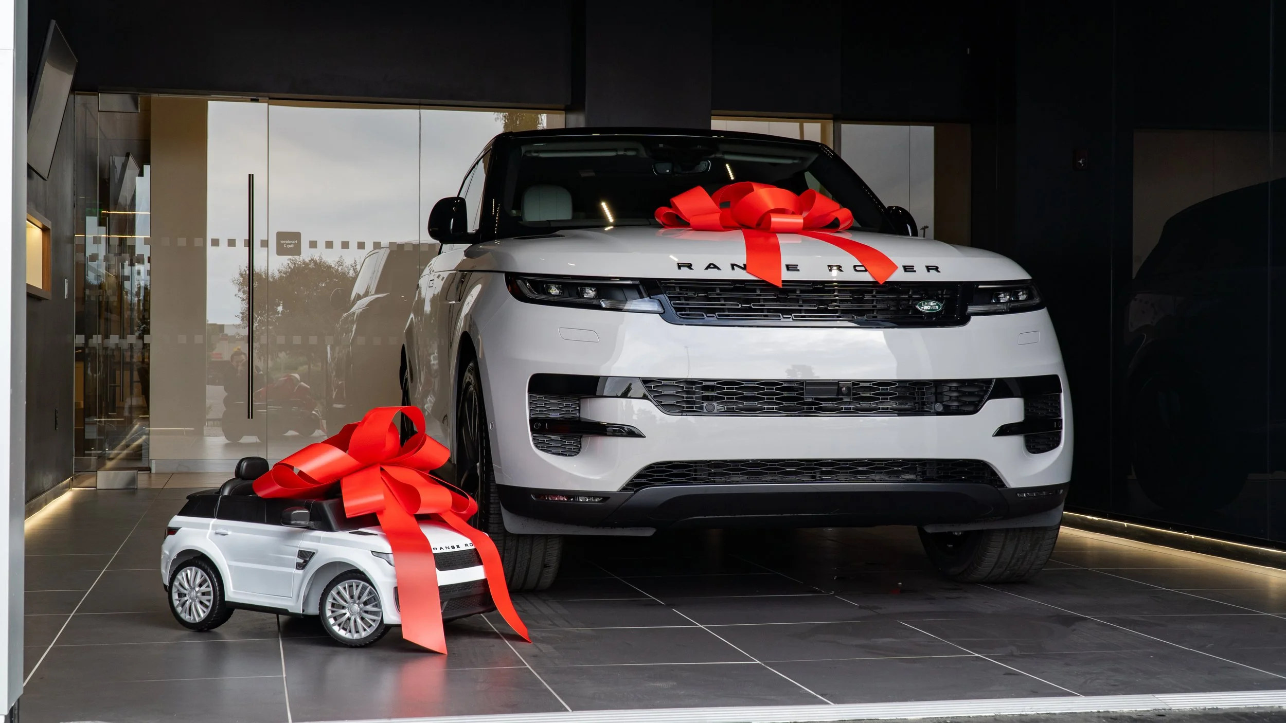 White Range Rover with a large red bow on its hood and a small toy car with a red bow in front, both are placed inside a car dealership.