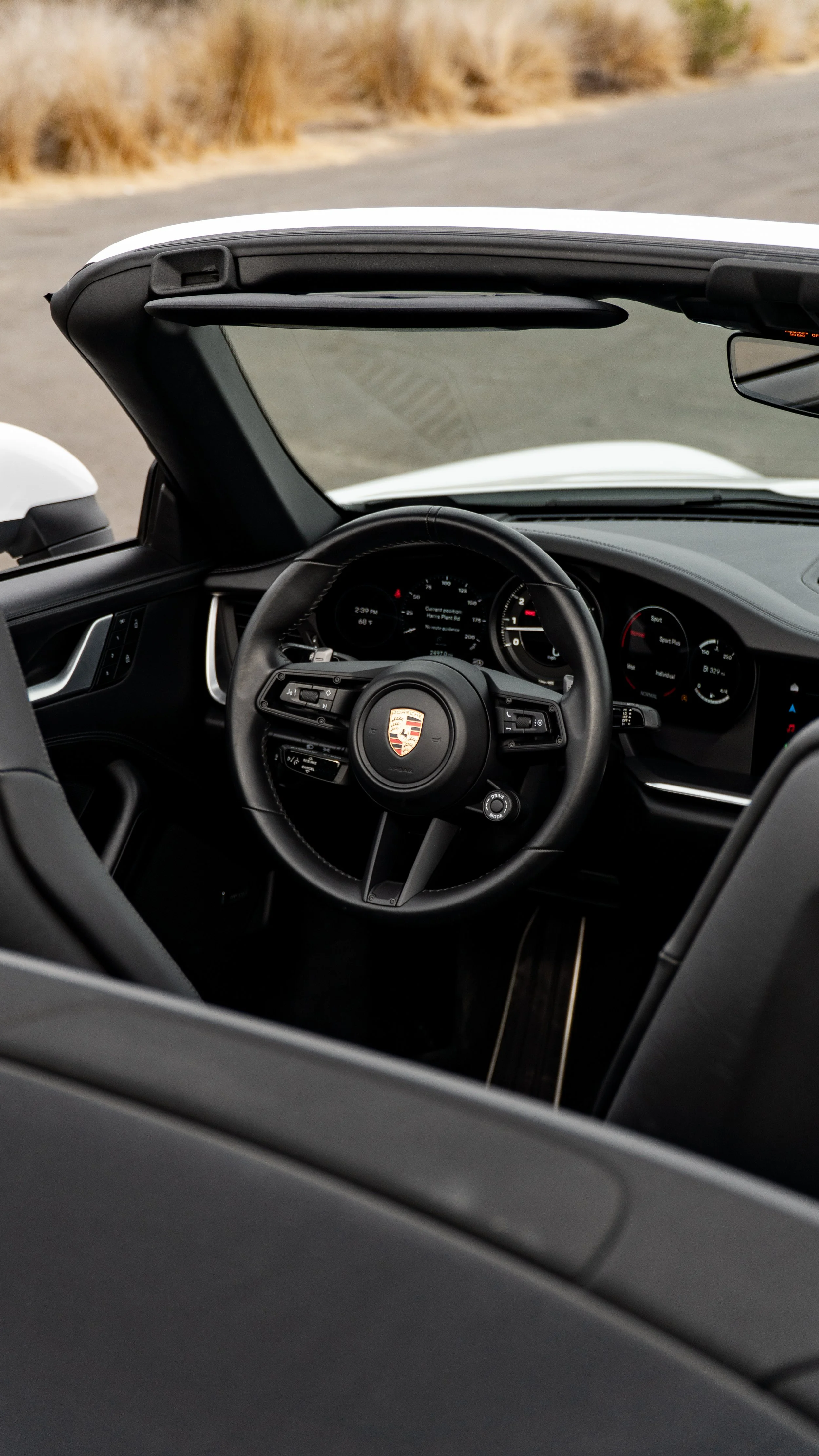 Interior view of a black Porsche convertible car showing the steering wheel with Porsche logo, dashboard, and part of the windshield, with desert landscape in the background.