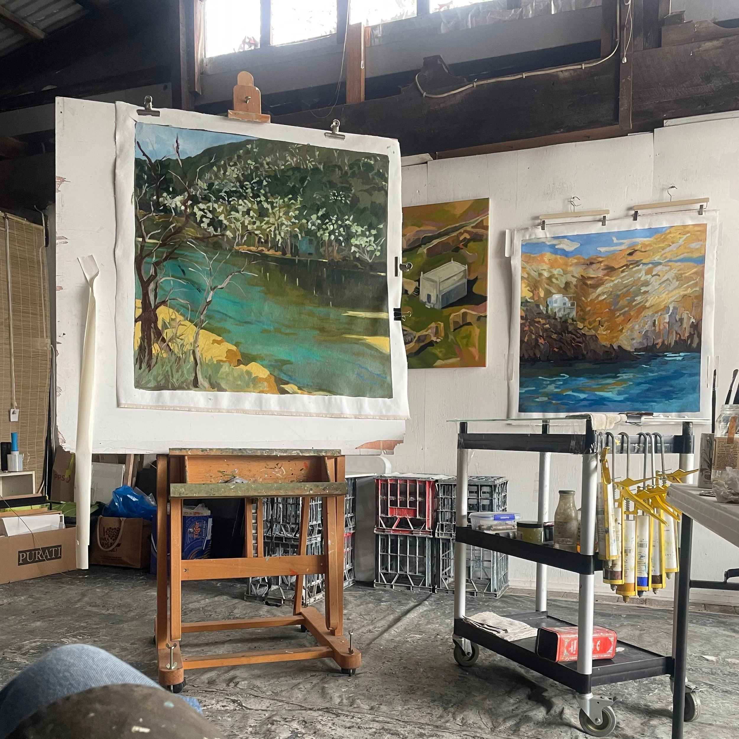 An art studio with three landscape paintings on the wall, an easel, a rolling cart, and various art supplies.