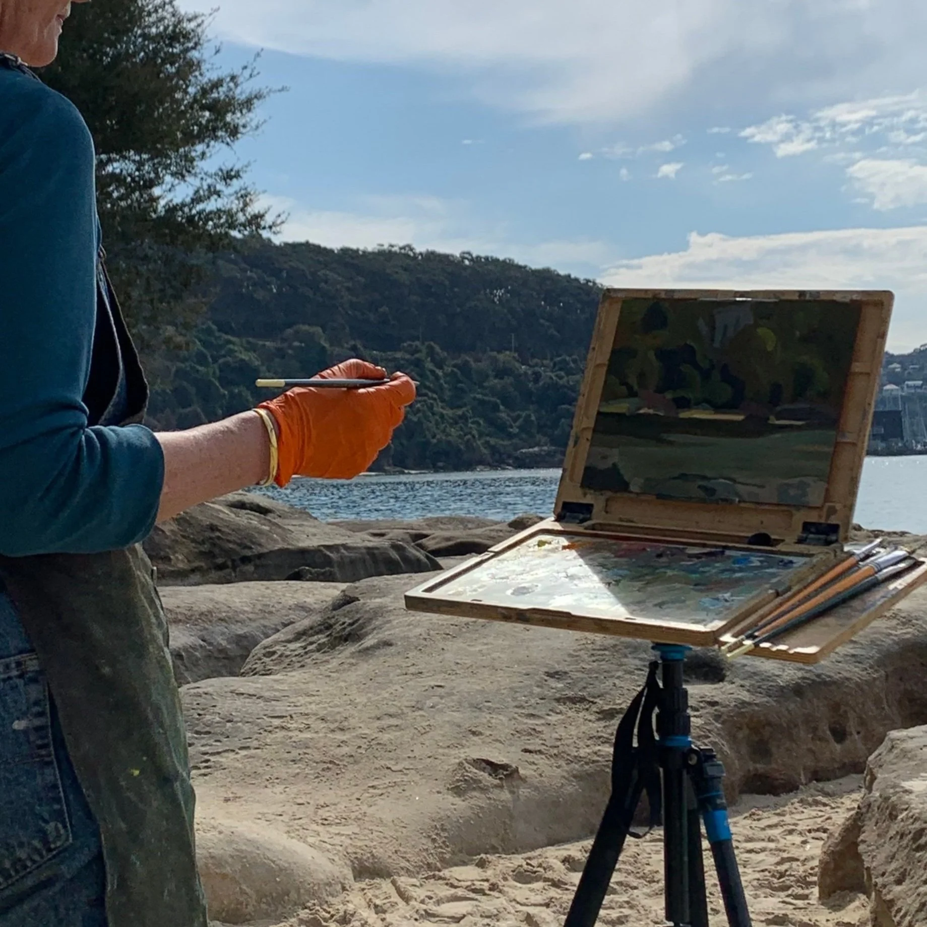 A person wearing an orange glove holding a paintbrush, standing on a rocky beach in front of an open painting set on a tripod. The painting depicts a landscape scene with trees and water.