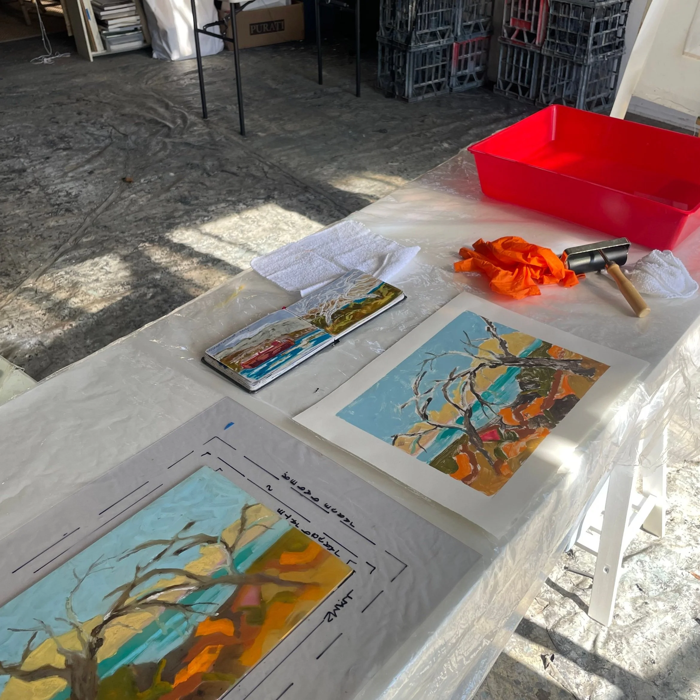 A table covered with a plastic sheet displaying two colorful paintings of trees, a small open photo album with painted artwork, an orange pair of gloves, a rubber mallet, a white towel, and a red tray. In the background, there are crates and boxes, and the floor appears to be concrete with natural light streaming in.