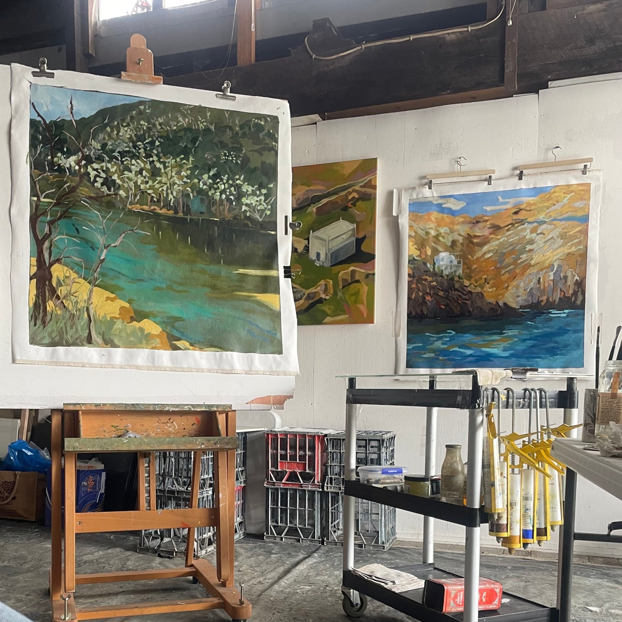 Art studio with three landscape paintings of nature scenes on the wall, and painting supplies on carts and crates.
