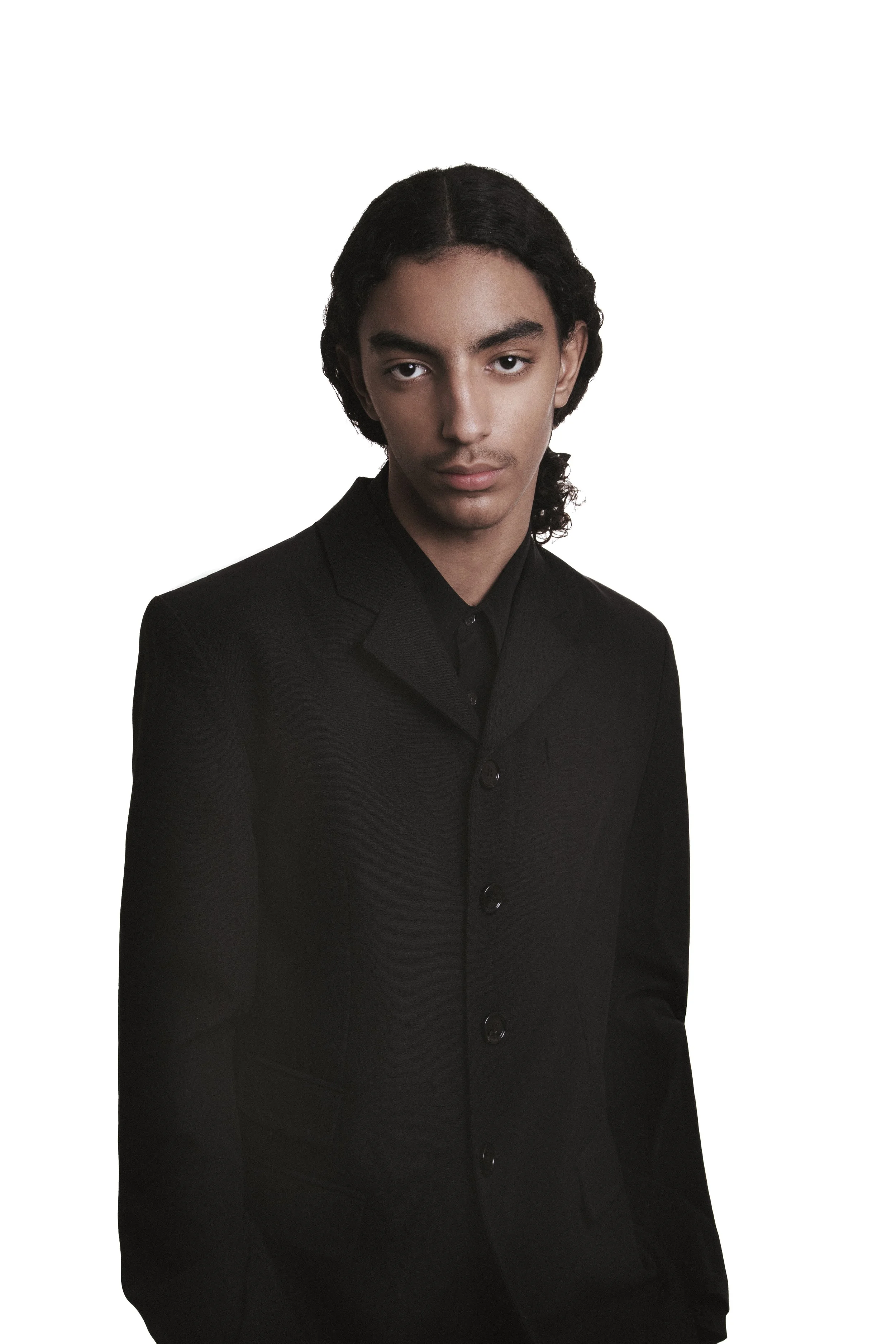 Jeune homme avec des cheveux longs, portant un costume noir, regardant sérieusement l'appareil photo, fond blanc.