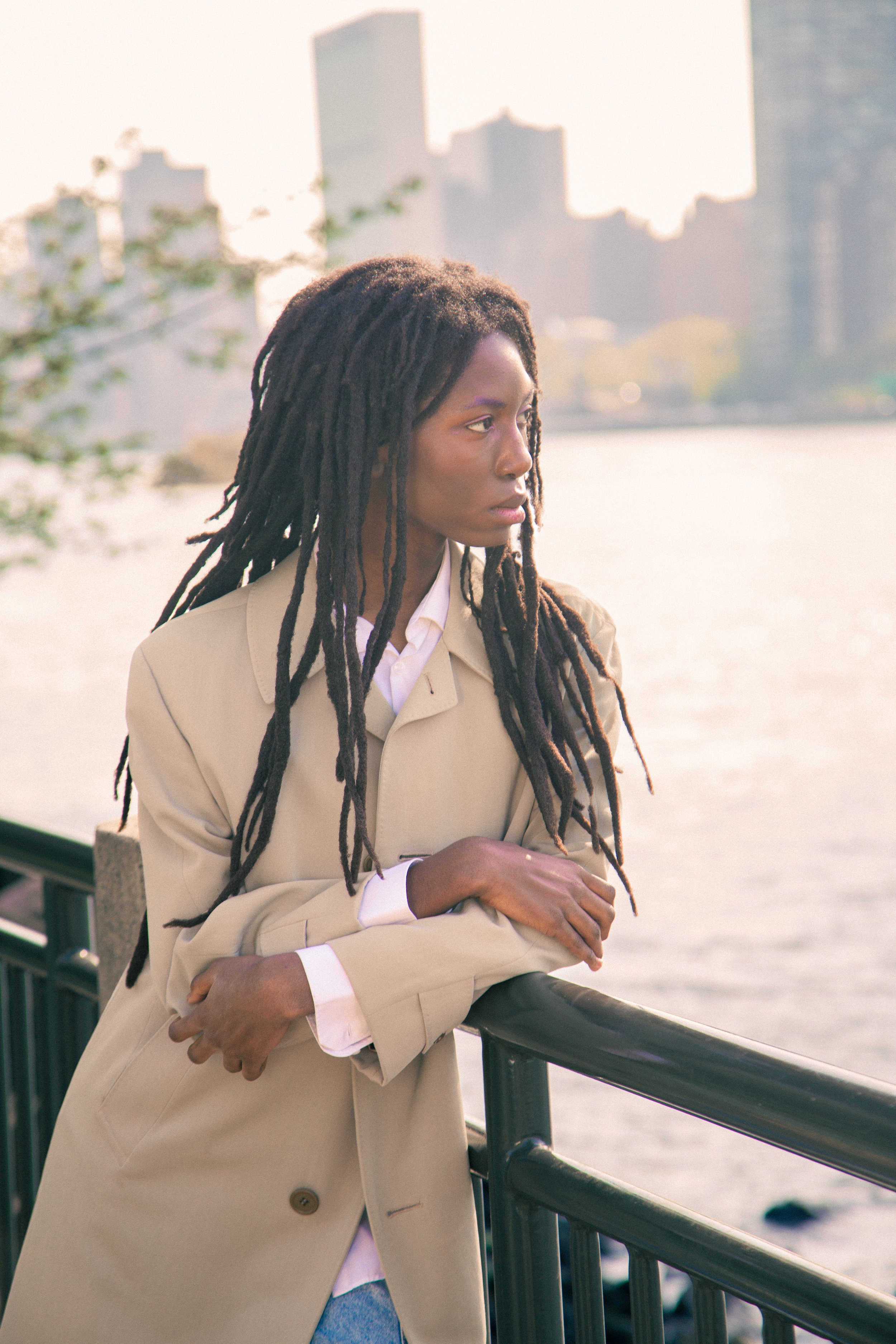 Femme aux dreadlocks regardant au loin, en costume beige, sur un quai au bord de l'eau, sur fond de skyline urbaine.