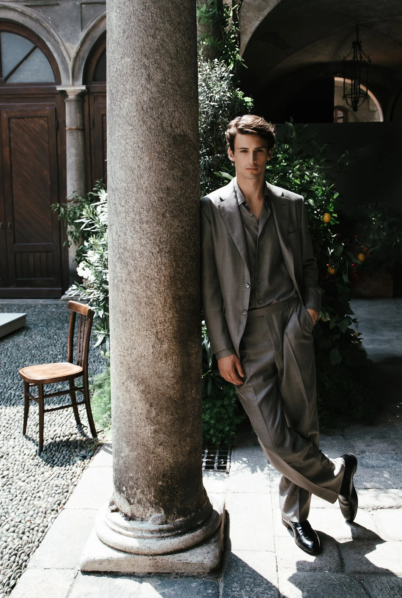 Jeune homme en costume gris décontracté debout près d'une colonne en pierre, dans un patio avec des plantes et une porte en bois en arrière-plan.