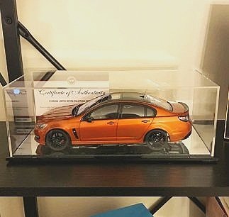 Orange model car displayed in a clear glass showcase on a black surface.