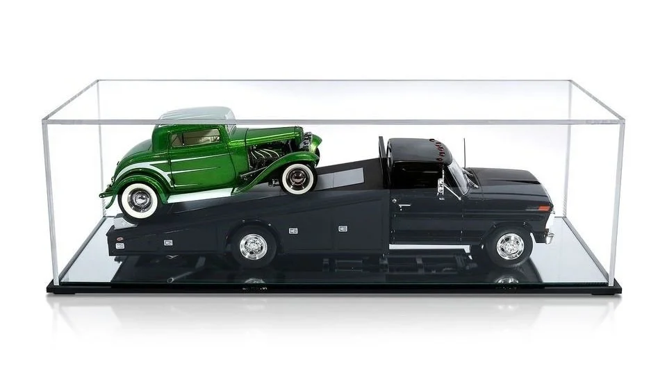 A model of a black pickup truck with a green vintage car on a raised platform inside a clear display case.
